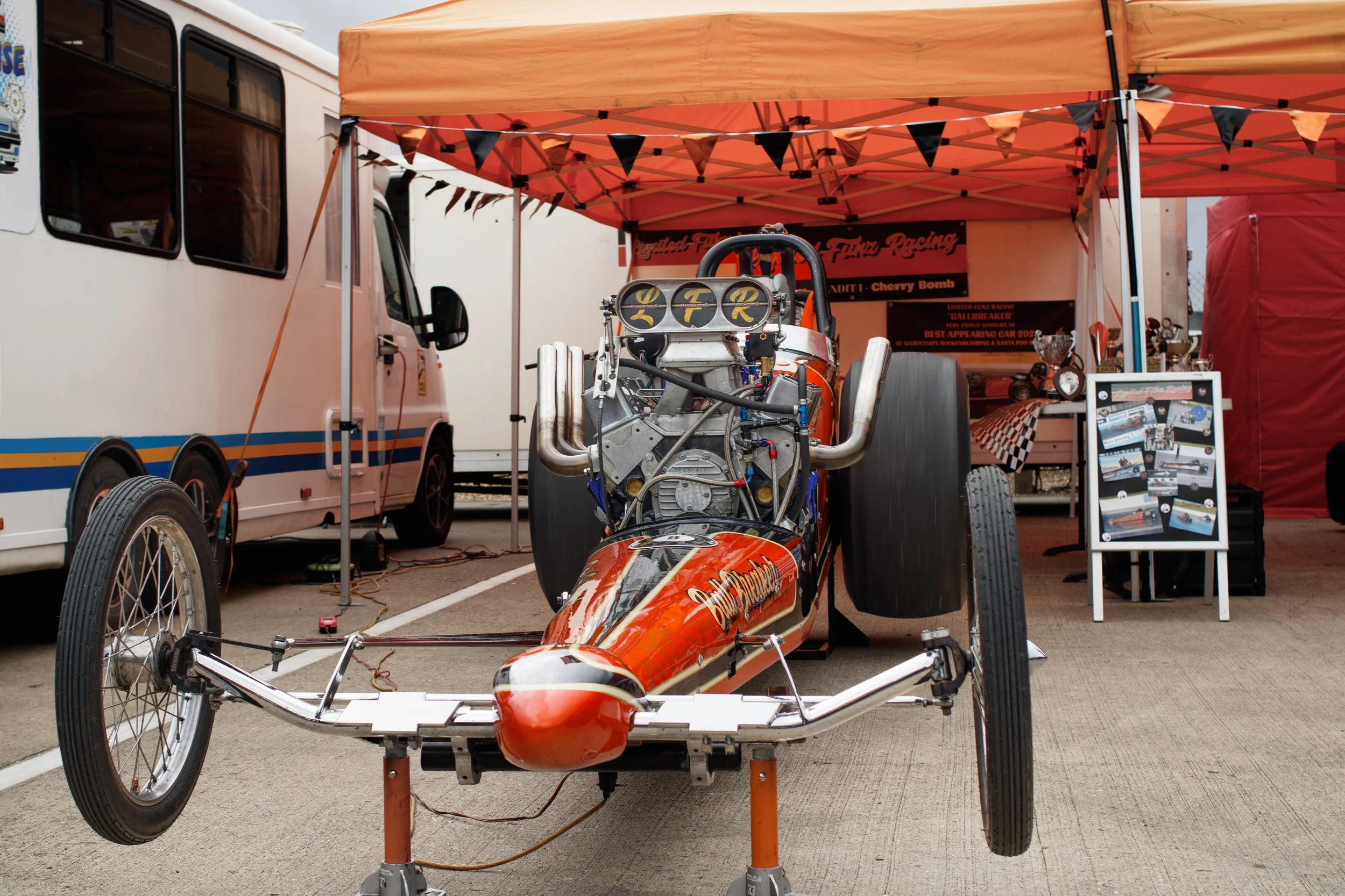 A vintage drag racing vehicle with an exposed engine, parked under a bright orange canopy at a racing event. The vehicle has large rear tires and a sleek, aerodynamic body with orange and black paint.