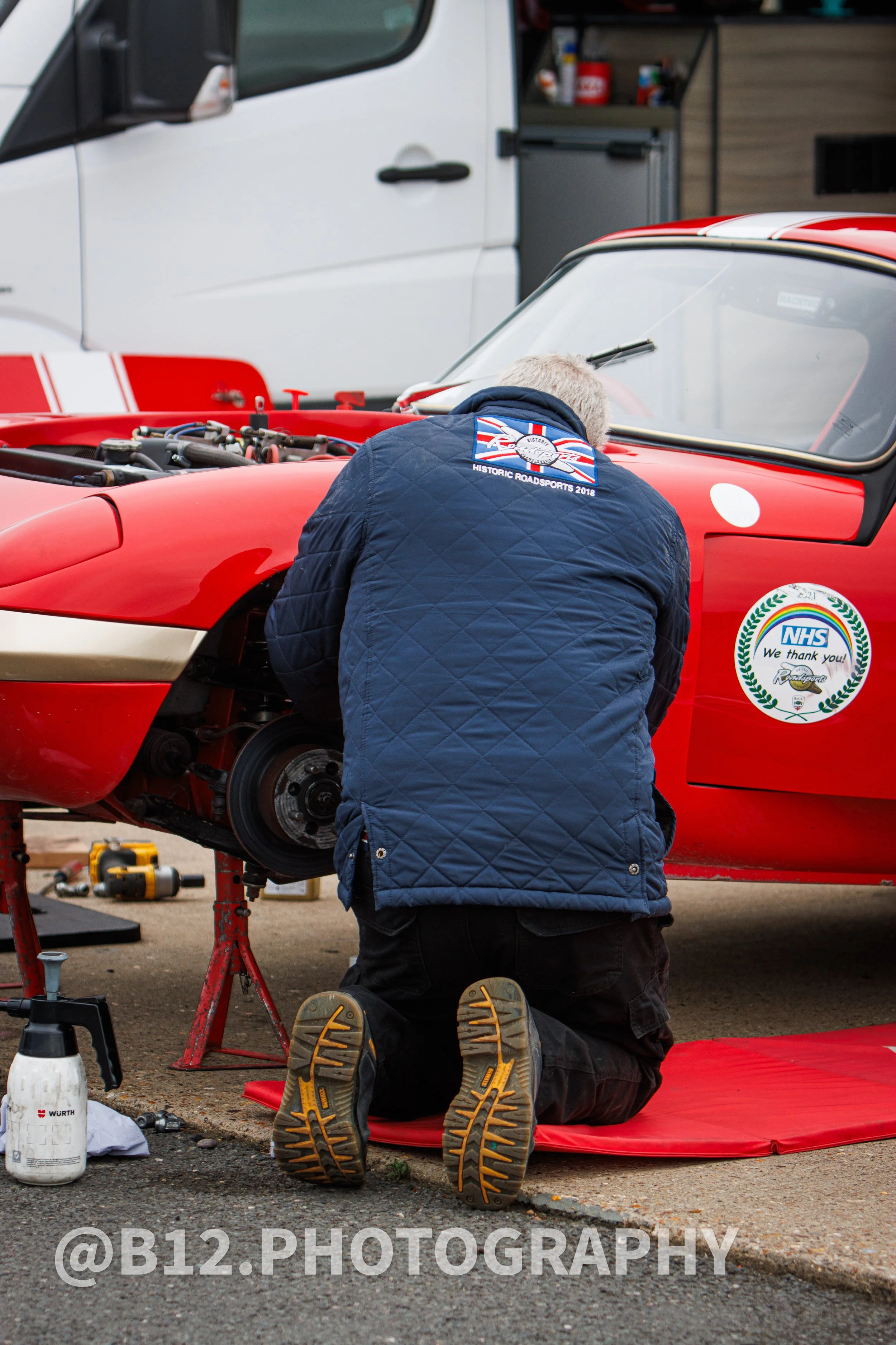 A man kneeling on a red mat, working on a red vintage race car with an open hood in a garage or pit area. The man is wearing a navy quilted jacket with a racing patch and has gray hair. Tools and a spray bottle are nearby.