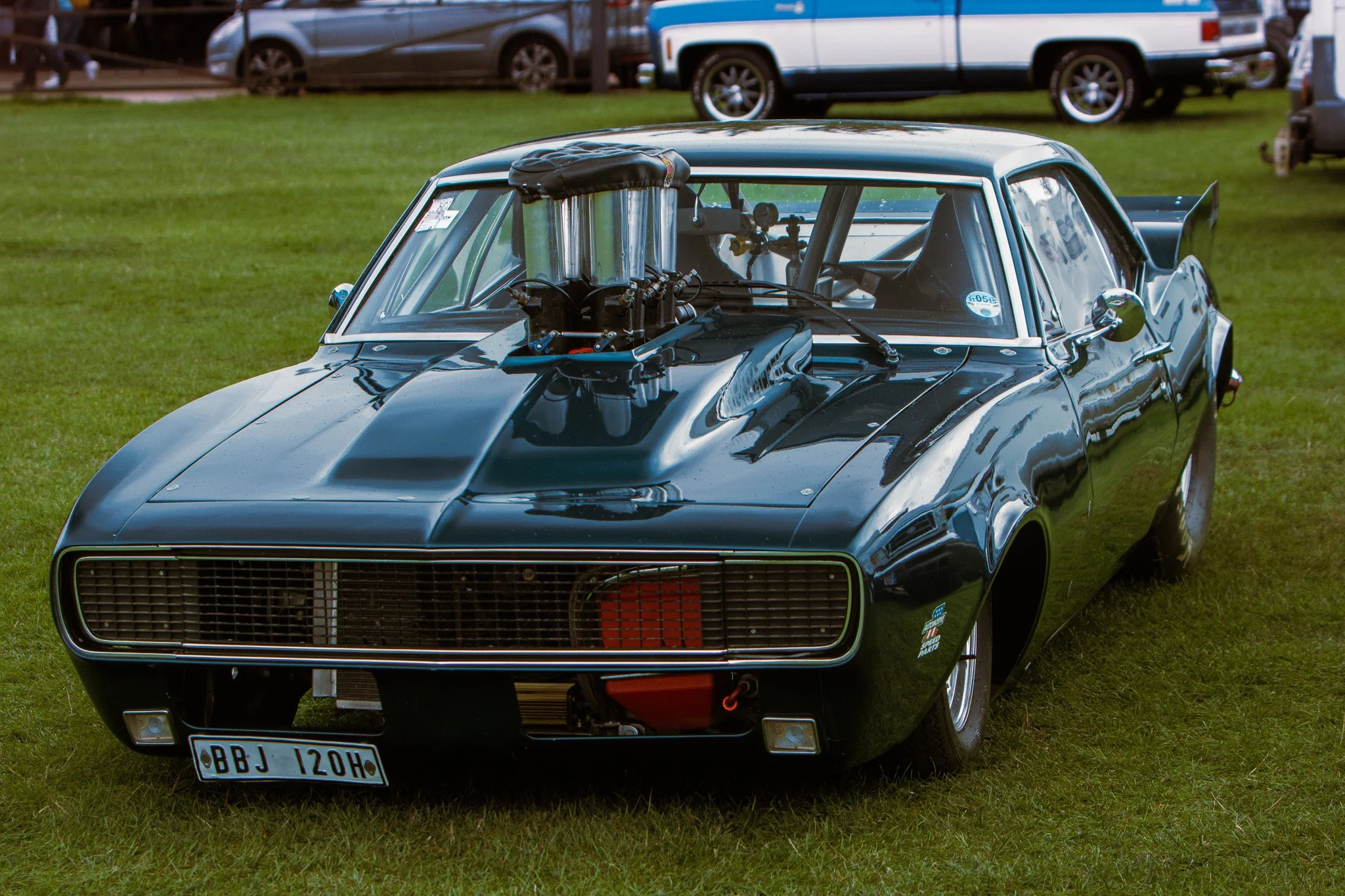 Black vintage race car with a supercharger on the hood, parked on a grass field at a car event.