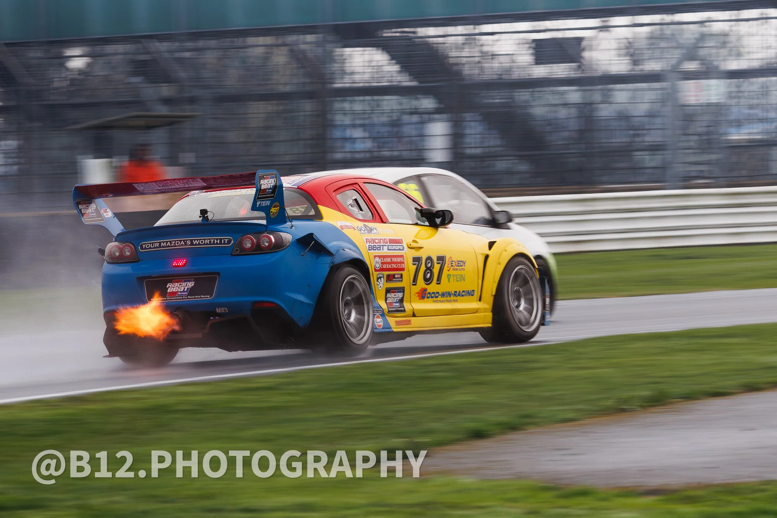 A blue and yellow race car with the number 787 on the side, speeding on a wet race track, with flames coming from the exhaust and a smoke trail behind it.