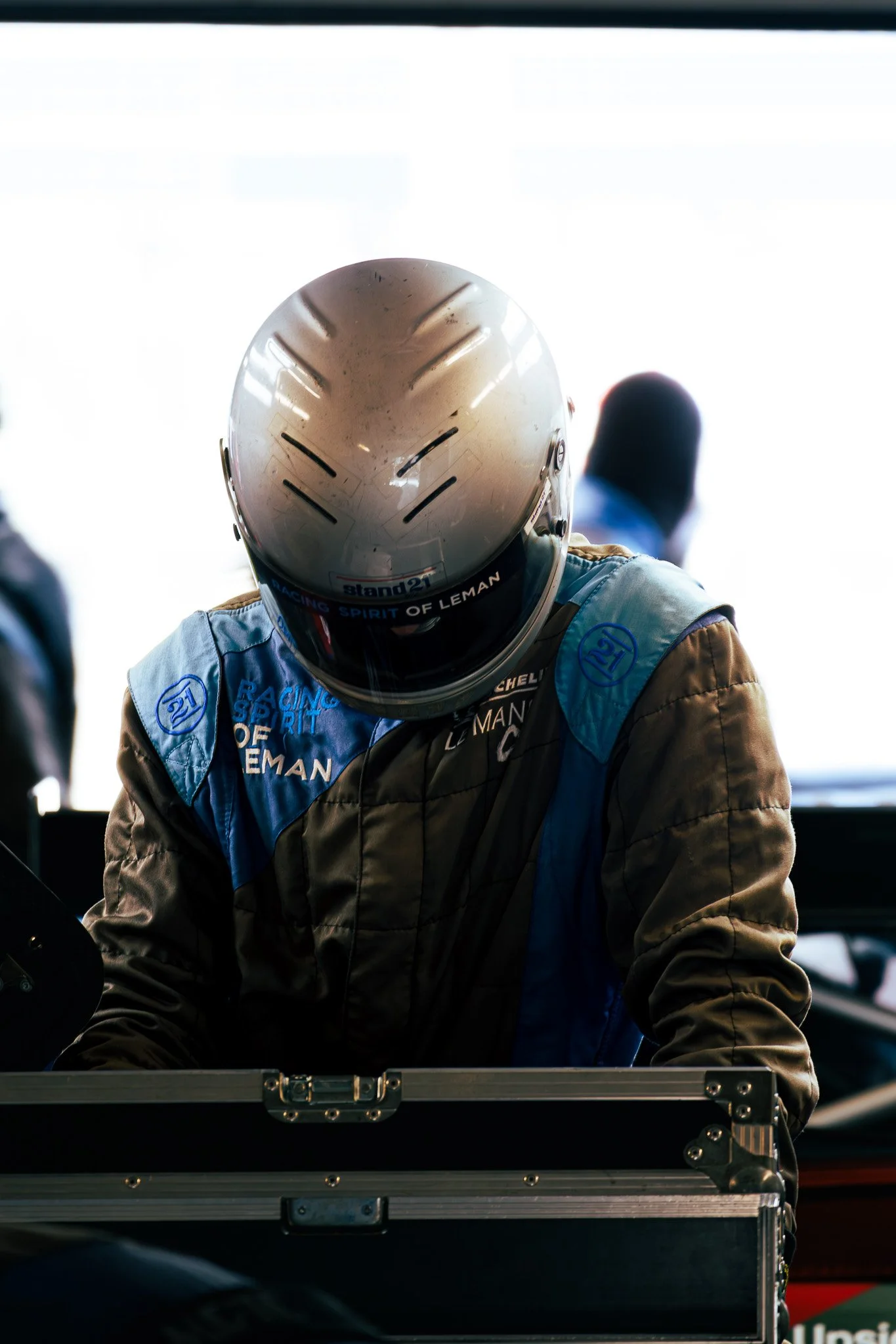 Race car driver in racing suit and helmet looking down, standing behind a case or equipment box, in a garage or pit area.