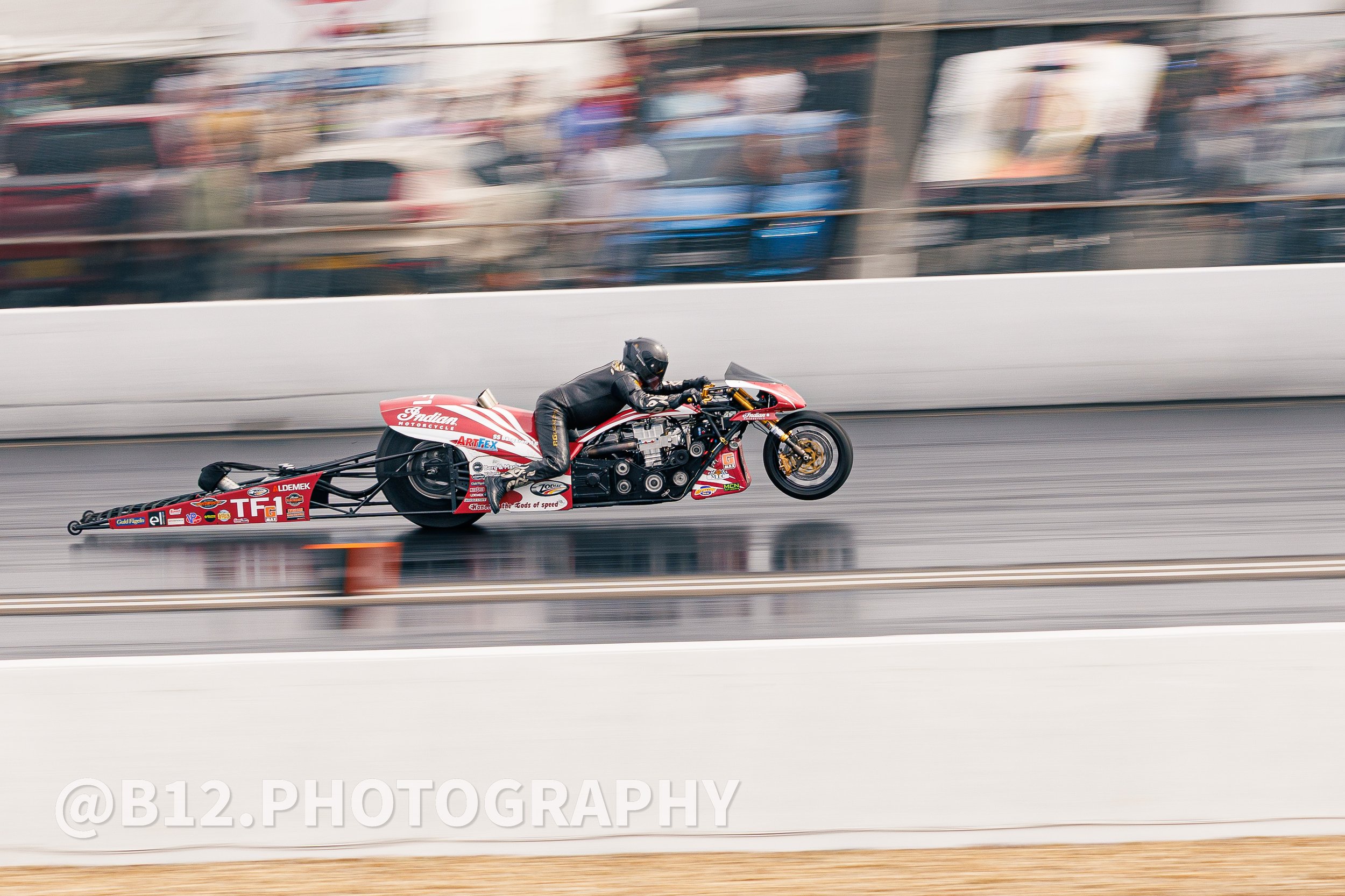 A motorcycle drag racing bike with a rider in black gear and helmet speeding down the track, with blurred background.