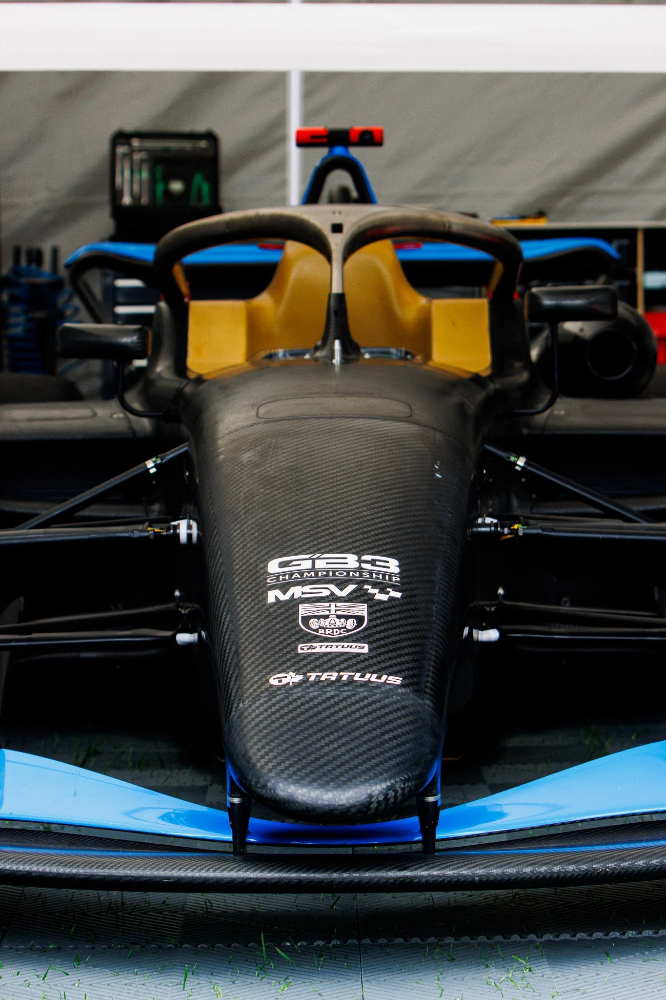 Close-up of a race car's front view, showing a carbon fiber body with various sponsorship stickers, in a garage setting.