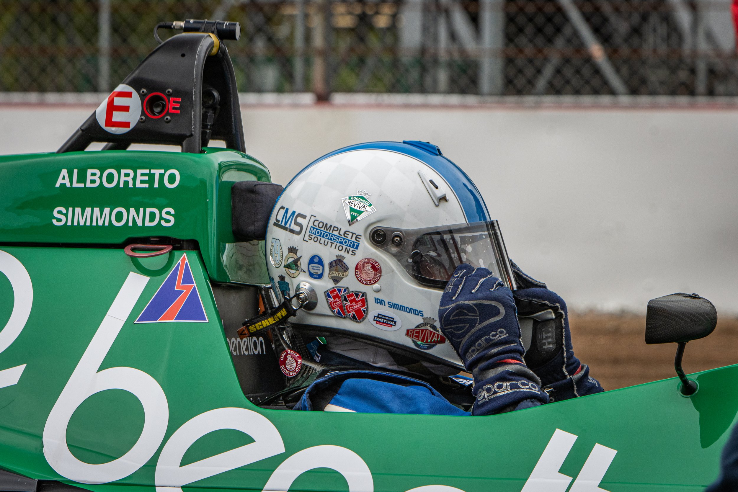 Race car driver wearing a white helmet with stickers, sitting in a green race car, adjusting their helmet.