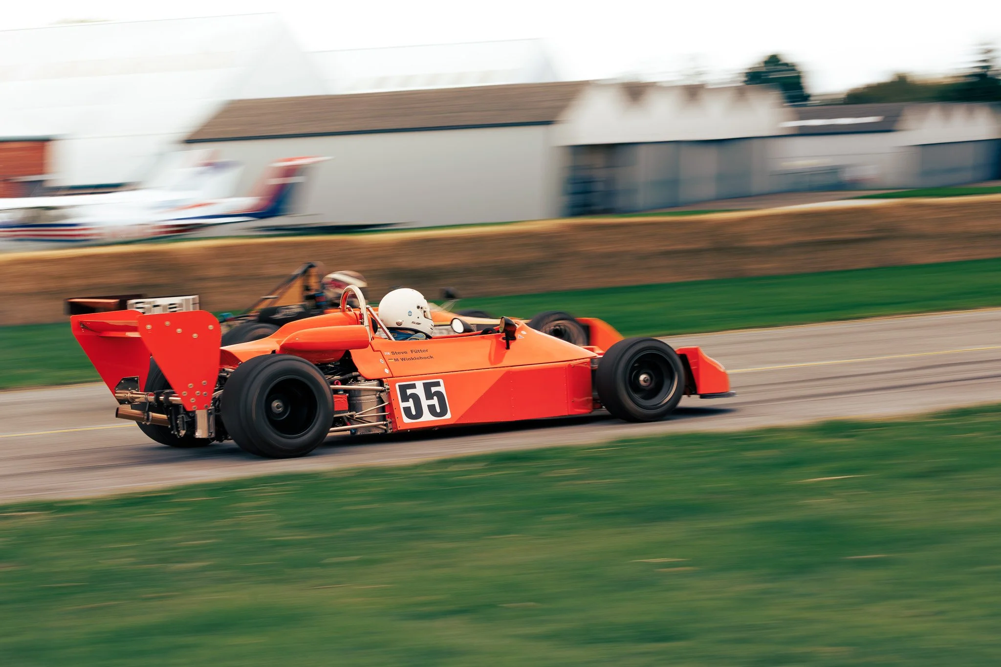 Orange vintage race car with number 55 on the side speeding on a race track, with a driver wearing a white helmet.