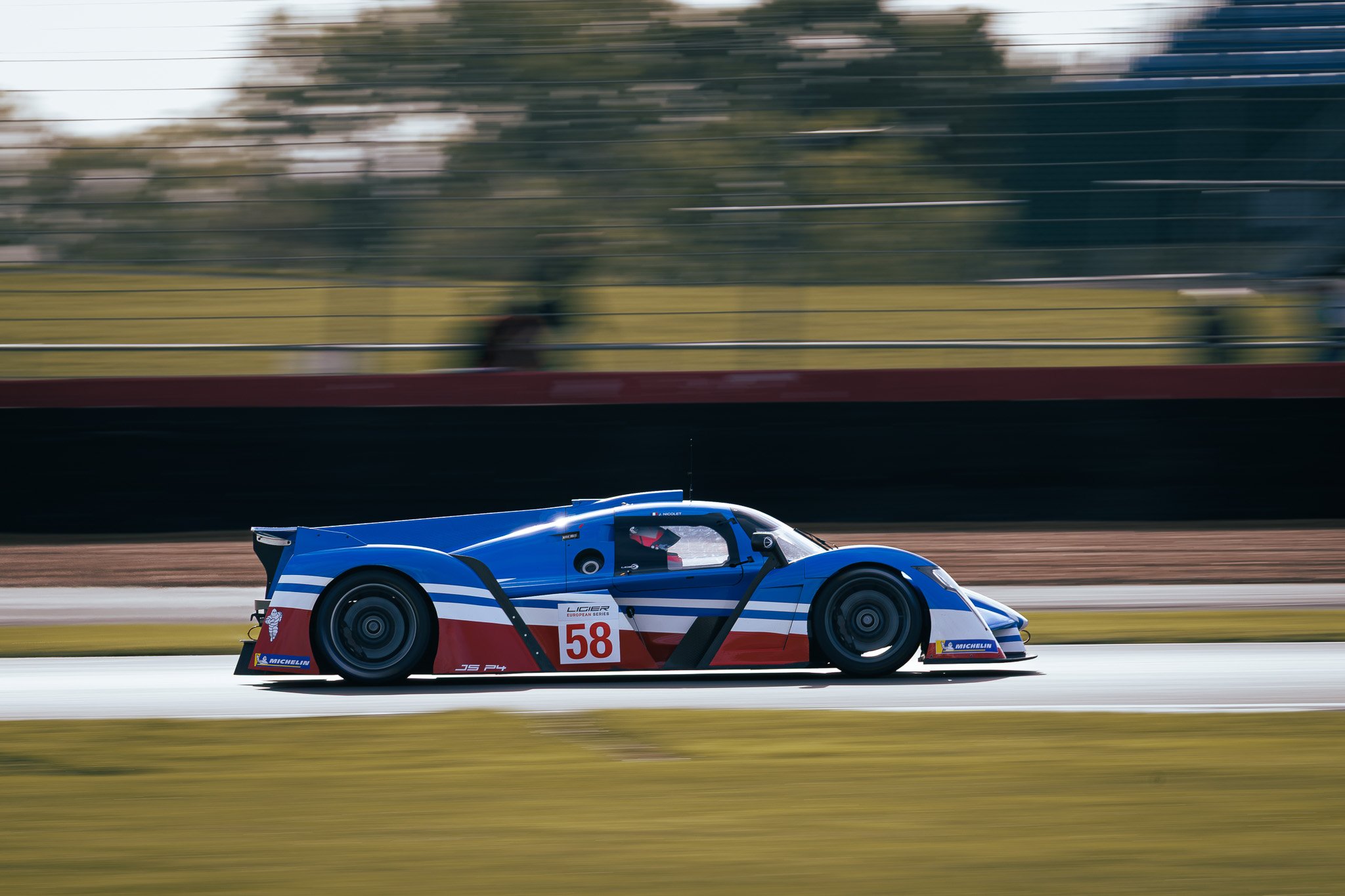 A sleek, blue racing car with red and white accents speeds along a racetrack during daytime, creating motion blur in the background.