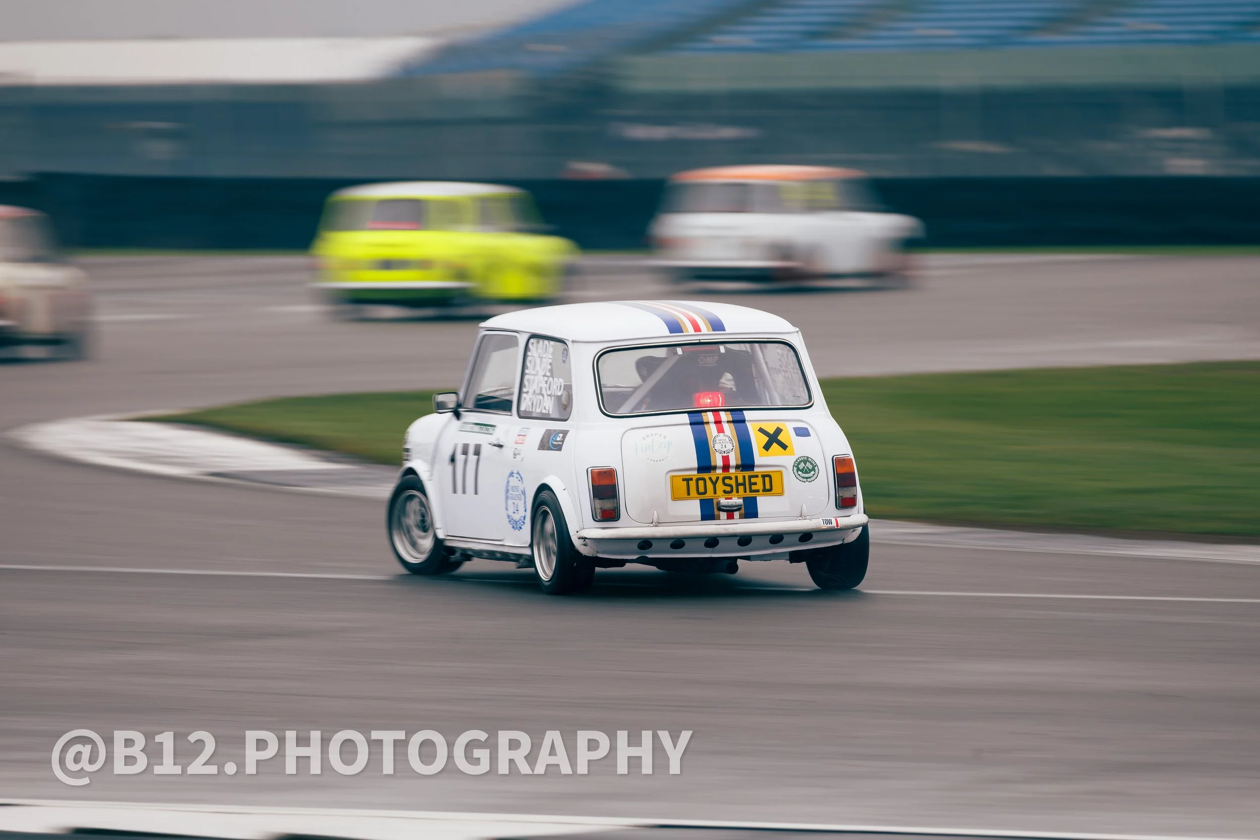A white vintage racing car with number 11 on its side and a 'TOYShed' license plate is speeding on a racetrack, with other cars in the background and motion blur indicating high speed.