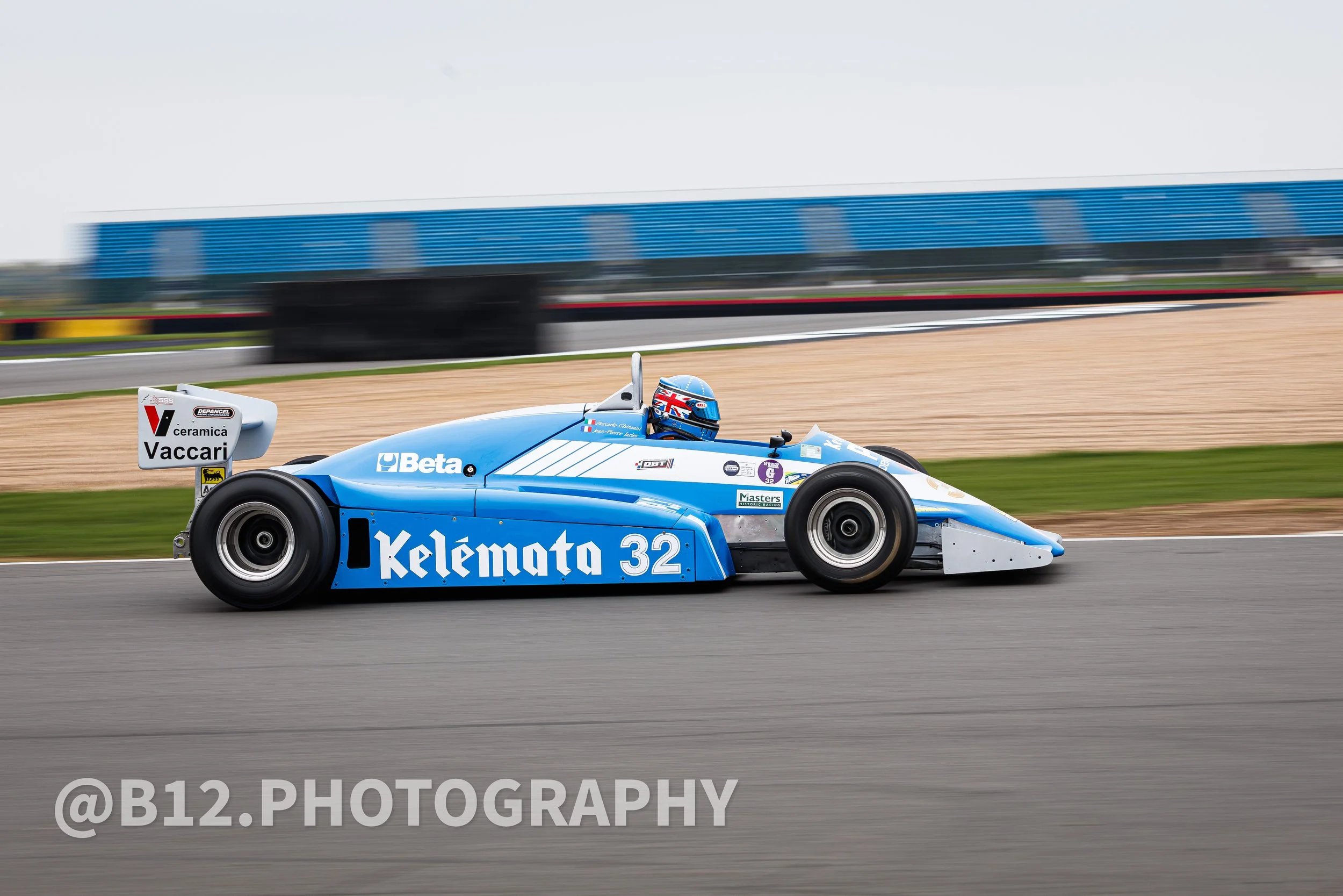 A blue vintage Formula racing car on a race track with a driver wearing a helmet with British flag design, advertising sponsors on the car, and empty grandstand in the background.