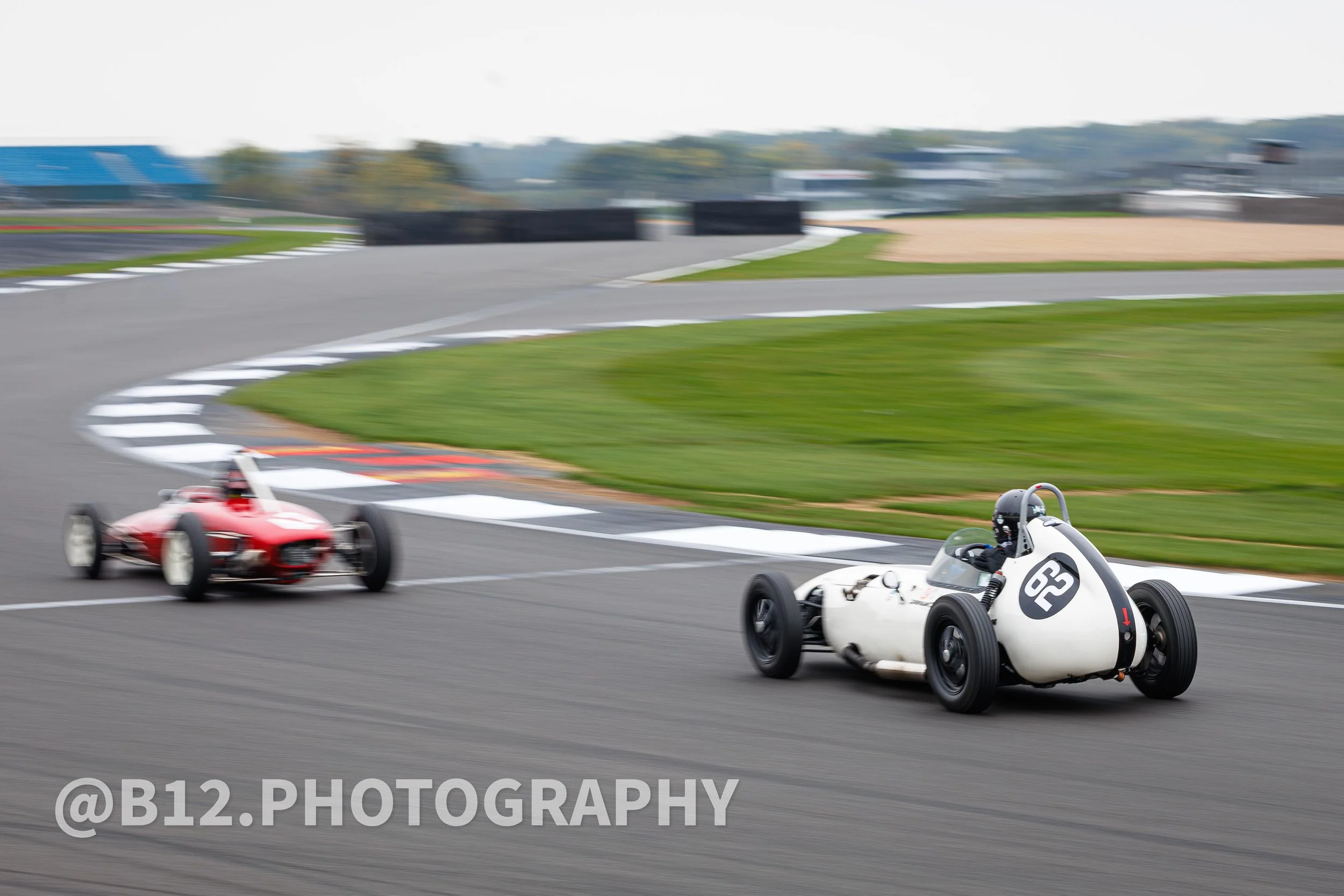 Two vintage race cars on a race track, one white with black number 62, and one red, speeding around a curve with blurred background.