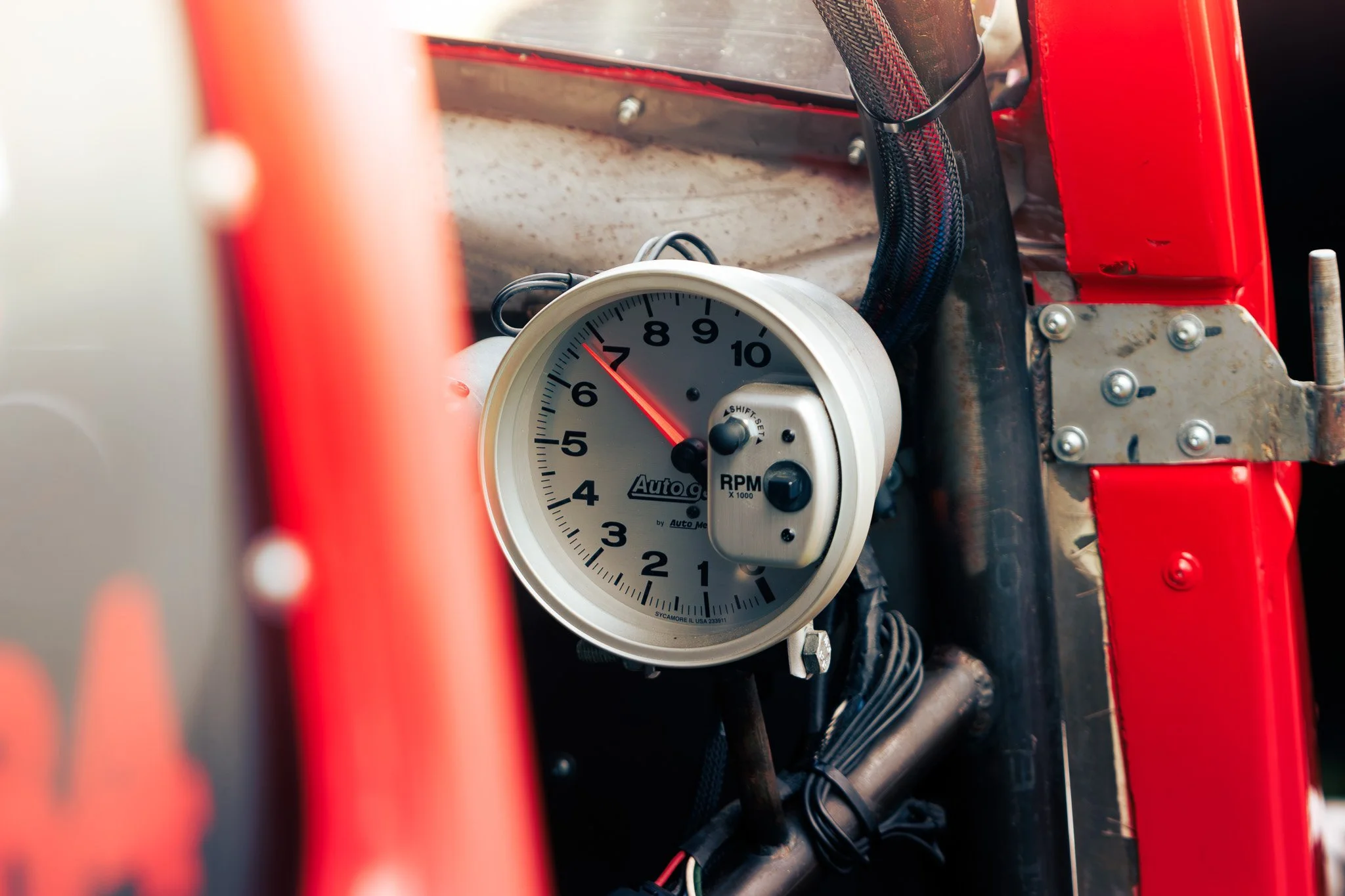 Close-up of a white tachometer with a needle pointing to 6, mounted inside a race car with red and black interior parts.