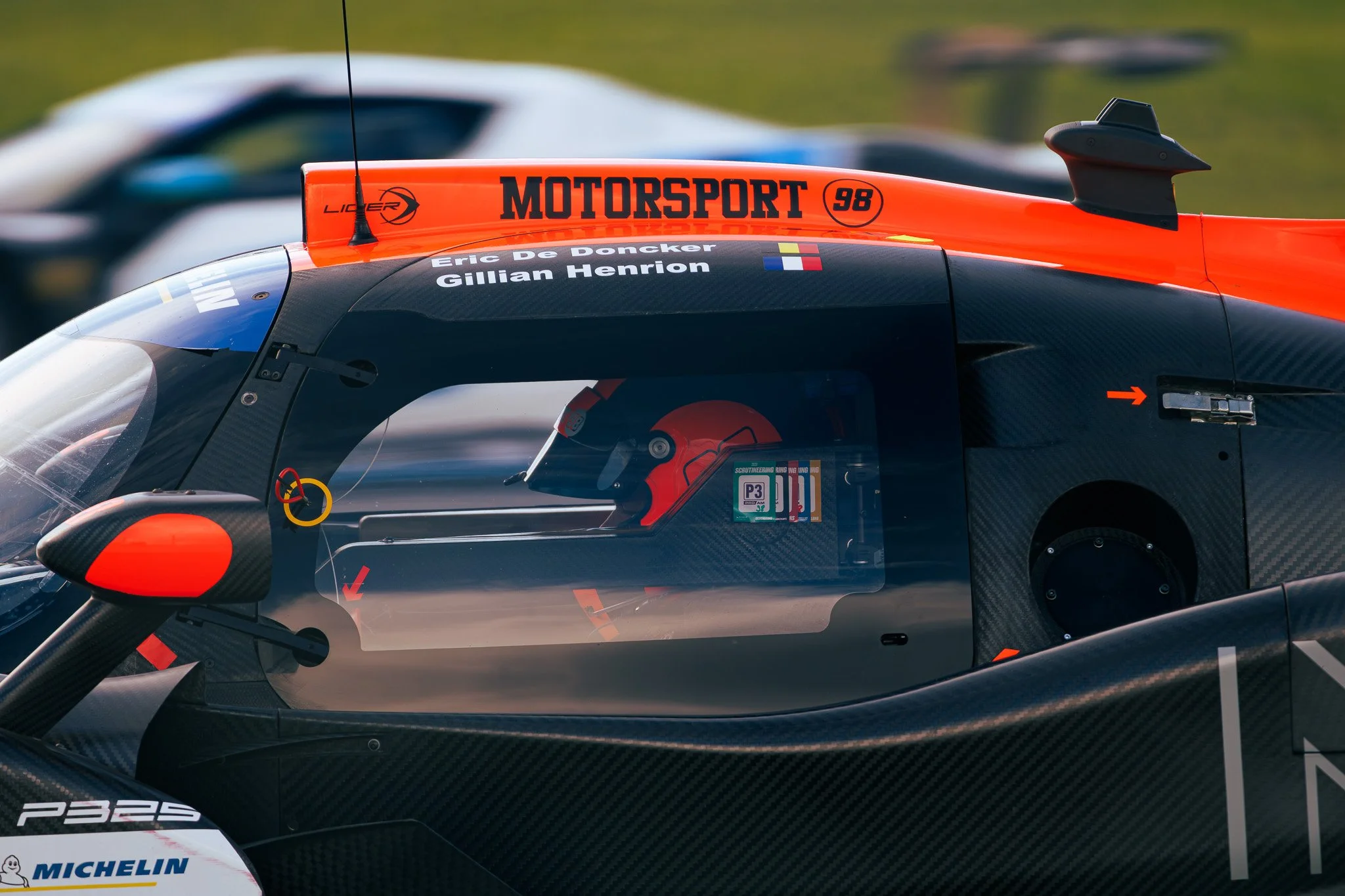 Close-up of a racing car cockpit with a driver wearing a red helmet, displaying racing decals and sponsors on the car's carbon fiber body, including 'MOTORSPORT' and 'Eric De Doncker'.