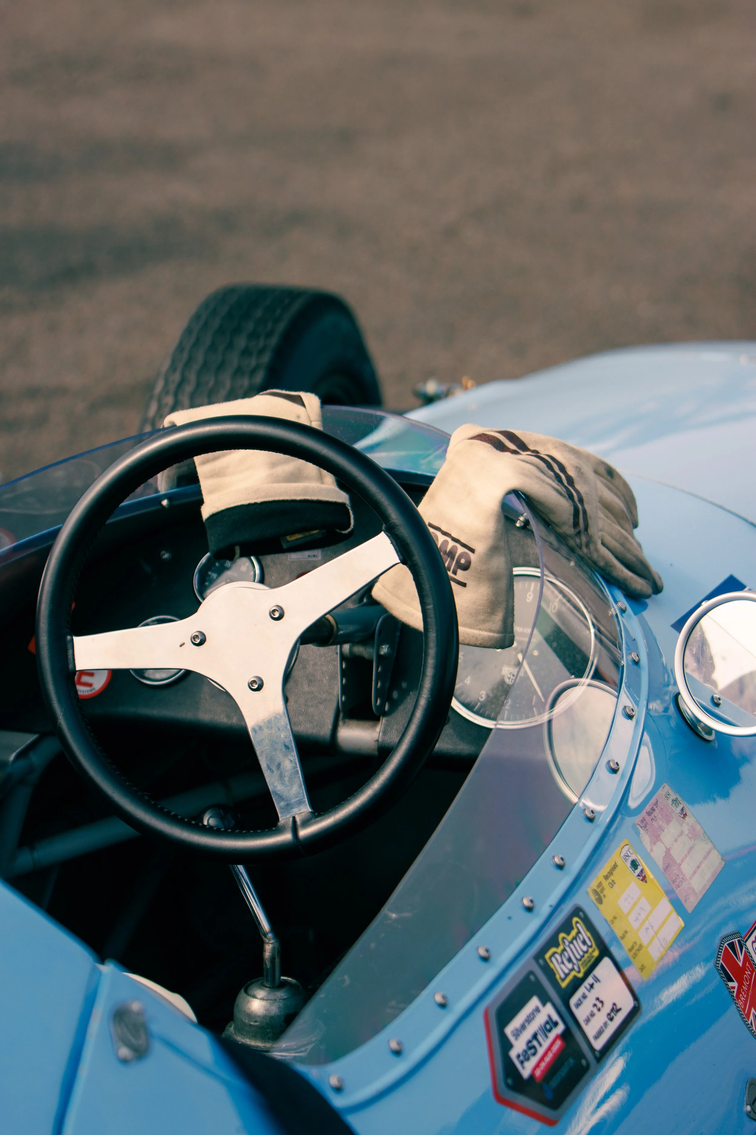 Close-up of a vintage race car dashboard with a steering wheel, racing gloves, and various stickers on the body.