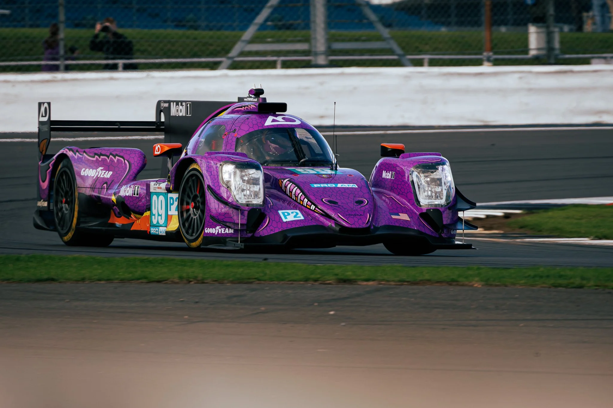 A purple race car speeding on a track, with a covered driver visible through the windshield, and spectator stands in the background.