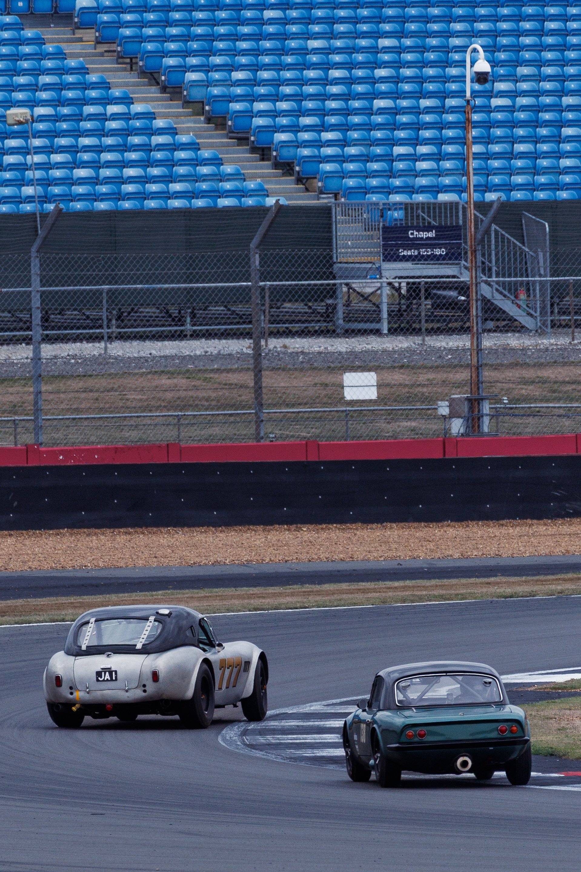 Two vintage race cars on a track, one white with the number 777 and a black roof, and the other green with a rear window, driving on a curved section of the raceway, with empty blue stadium seats and a chain-link fence in the background.