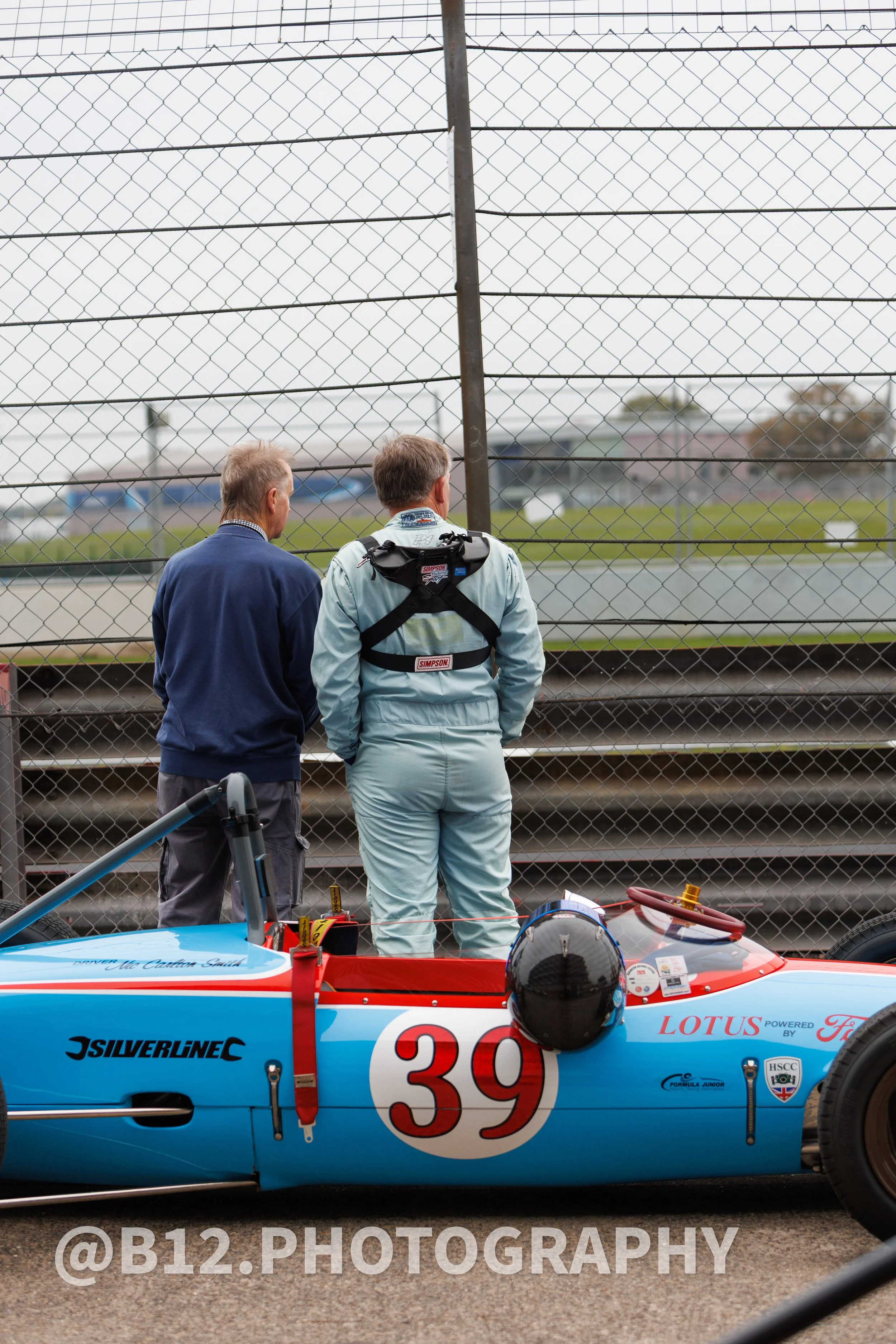 Two men stand at the race track, one wearing a racing suit and helmet, next to a vintage Lotus race car numbered 39, behind a chain-link fence.