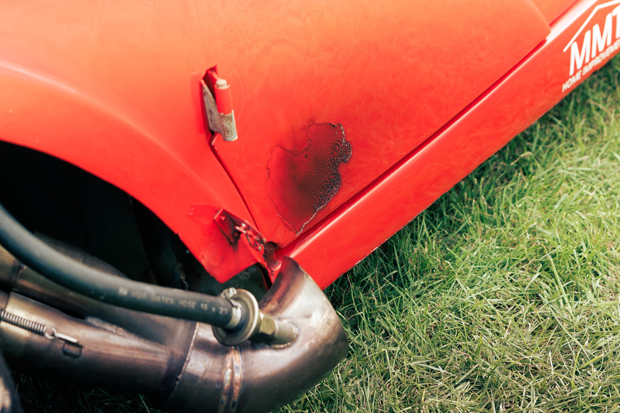 Close-up of a red race car's damaged and burnt side panel near the wheel, with visible black burn marks and scuffed paint, on grass.