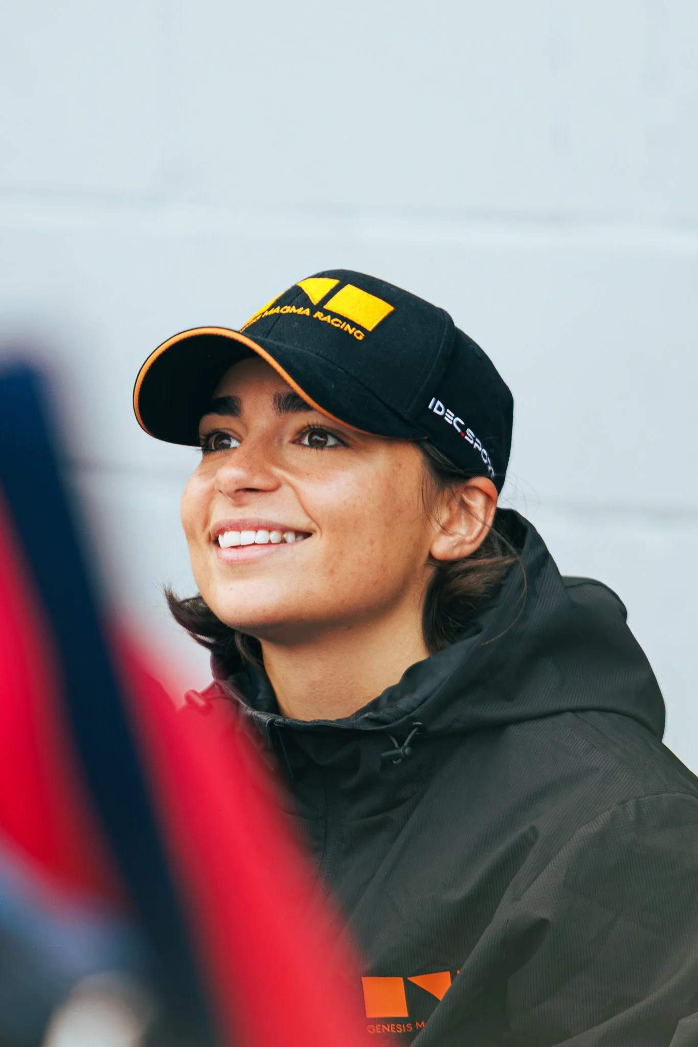A woman wearing a black cap with yellow and white logos and a black jacket, smiling and looking upward against a blurred background.
