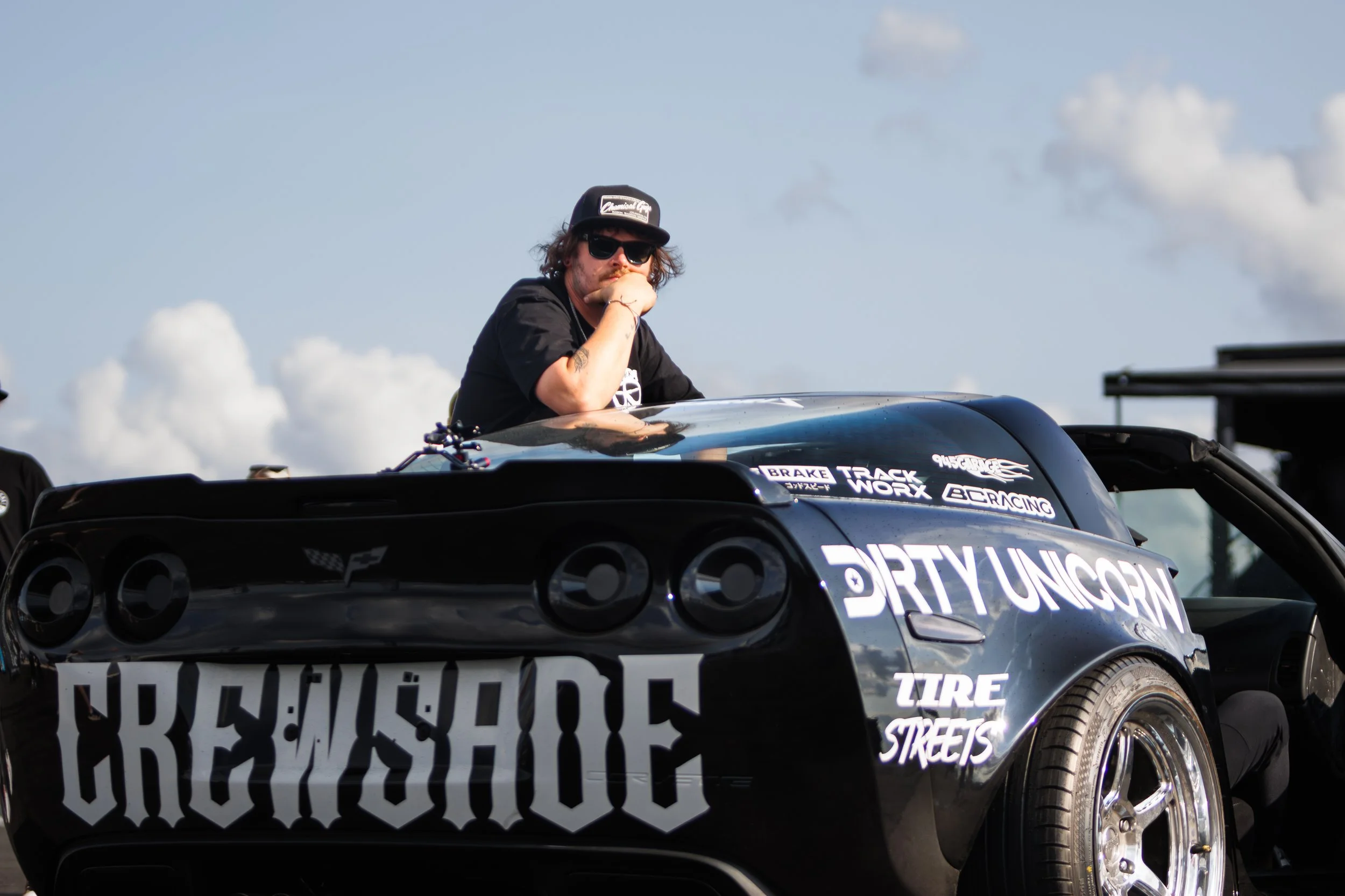 A man with sunglasses and a cap sitting on the hood of a black race car with various sponsor logos and decals, including 'CREW SHOE,' 'DIRTY UNICORN,' and 'TIRE STREETS,' looking over the scene with a contemplative expression. The sky is partly cloud