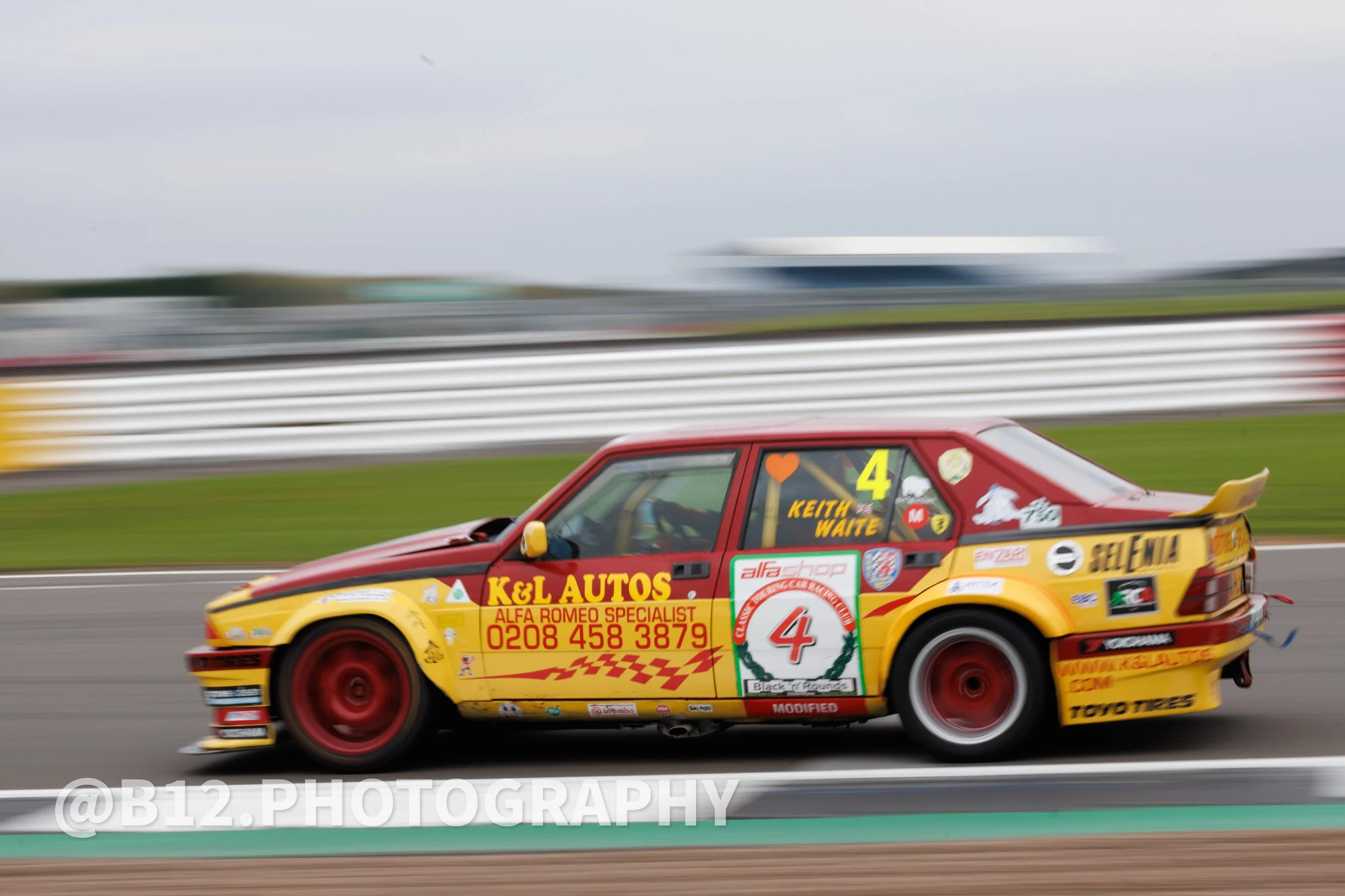 A racing car on a track with a red and yellow color scheme, sponsored by K&L Autos, with the number 4, driven by Keith Waite. The car is in motion, with a blurred background and wheels.