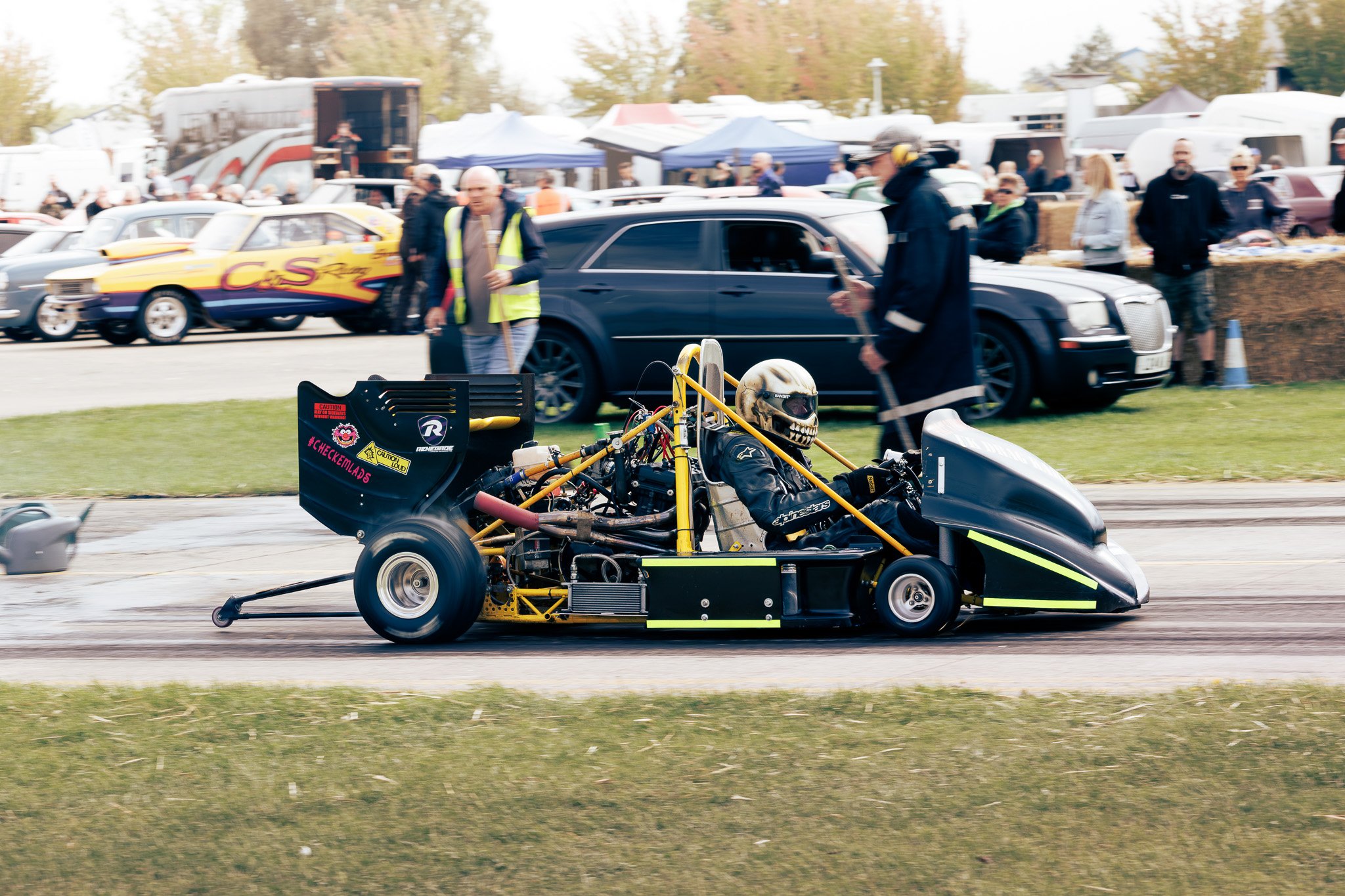 A go-kart race car speeding on a track with a driver wearing a skull helmet. The background shows spectators, vehicles, and tents at a racing event.