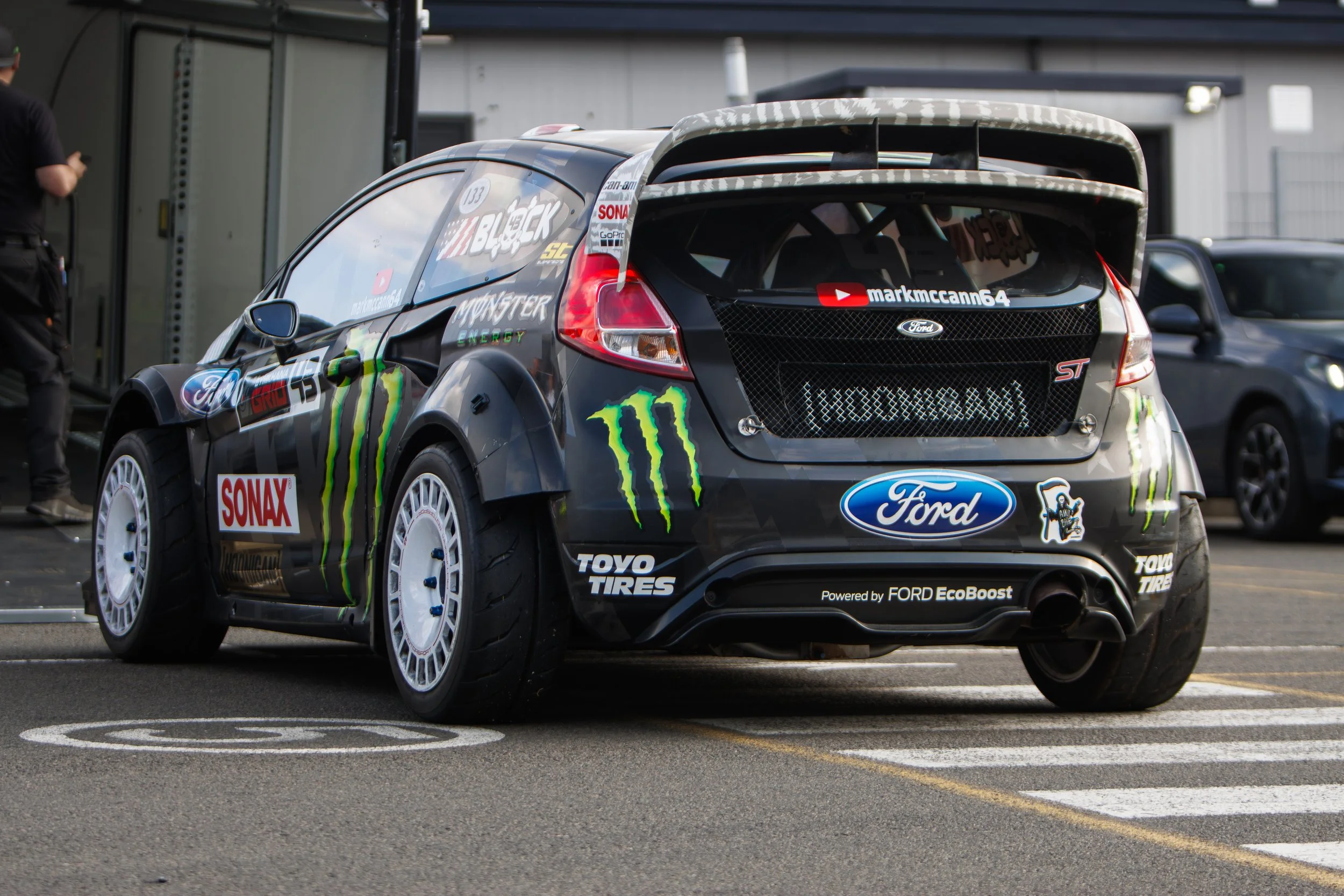 A black racing car with various sponsor logos, including Monster Energy, Ford, and Toyo Tires, parked in the paddock area. The car features a prominent rear wing and racing tires.