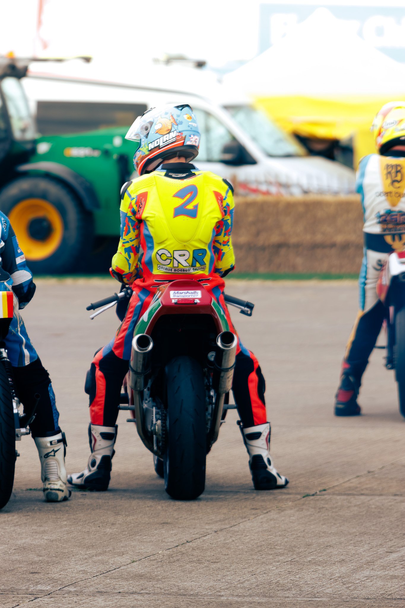 Motocross rider sitting on a motorcycle with their back facing the camera, wearing yellow and colorful racing gear and a helmet, on a racing track with other riders and vehicles in the background.