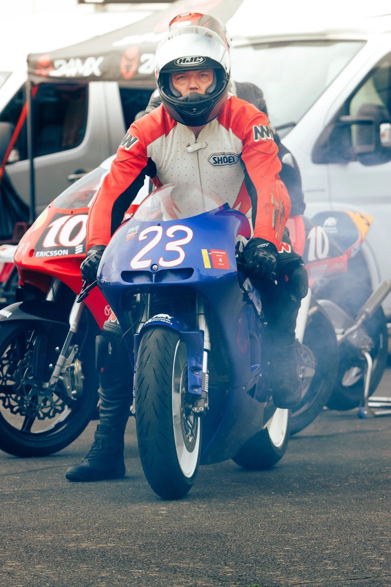 Motorcycle racer in a white and red suit sitting on a blue race bike with the number 23, holding the handlebars, in a racing paddock area.