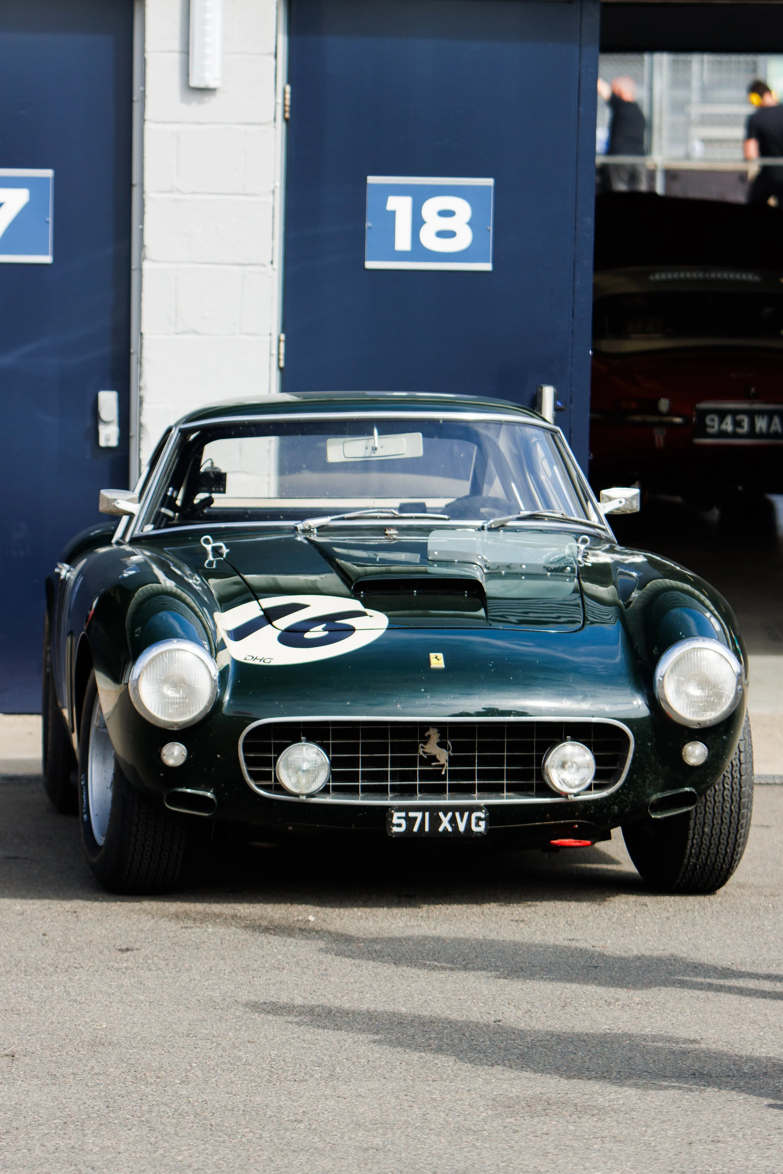 A classic dark green racing car with the number 76 on the hood, parked outside a garage with blue doors numbered 7 and 18. The car features round headlights, a Ferrari badge, and a vintage license plate 571 XVG.