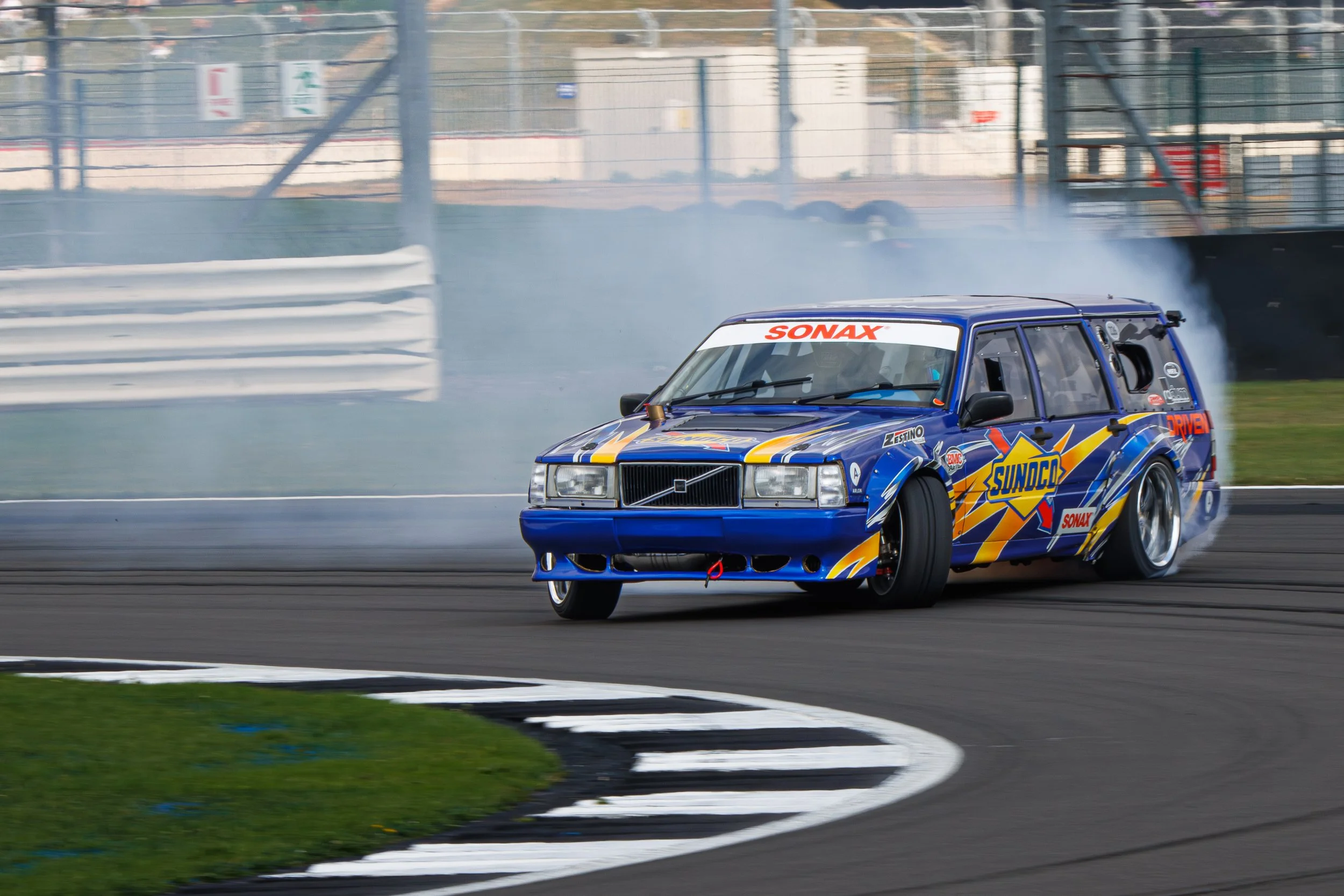 A race car drifting around a corner on a race track, with smoke coming from the tires, blue and yellow sponsor decals, and a white canopy with red text.