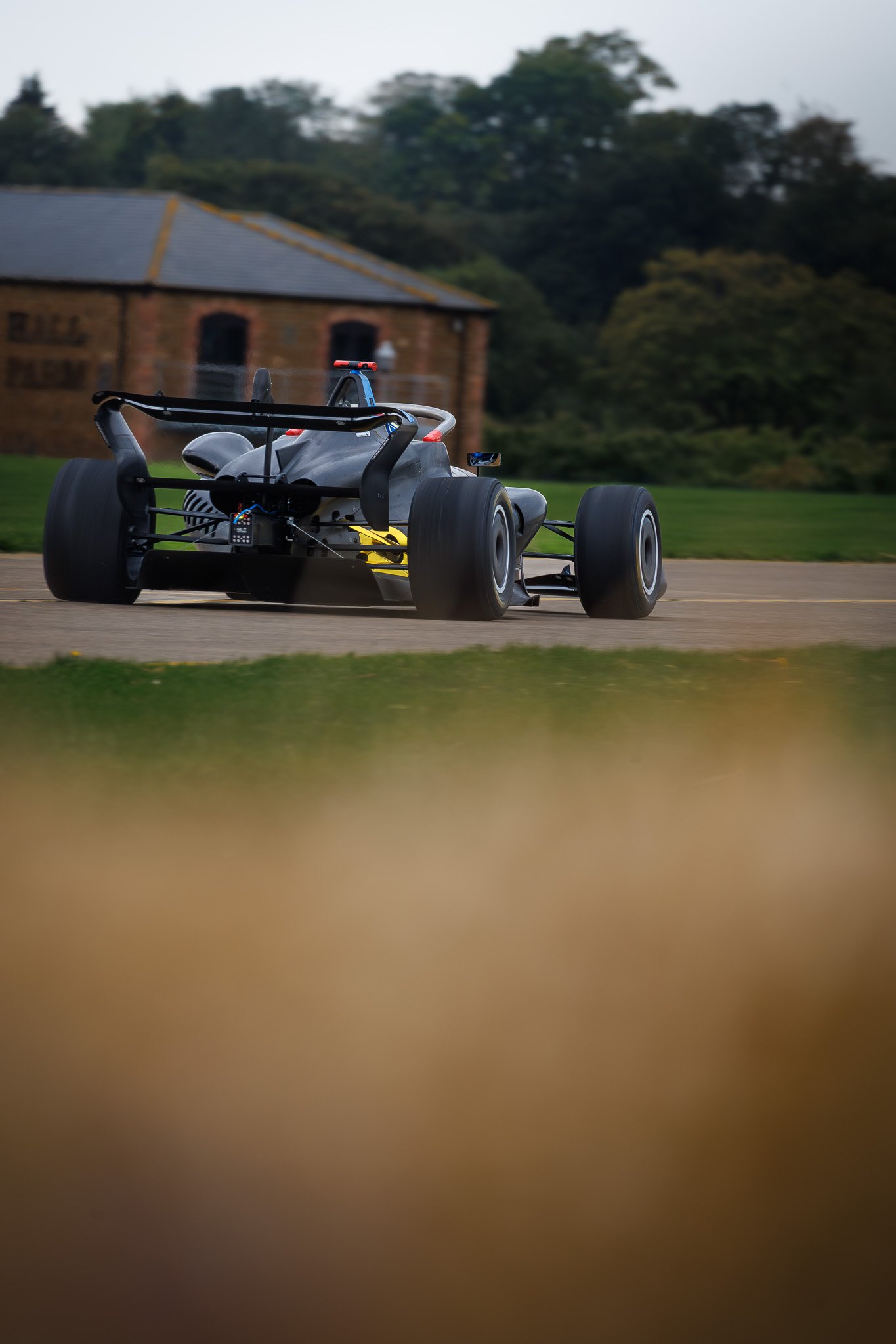 A sleek, gray race car speeding on a track, with greenery and a small brick building in the background.