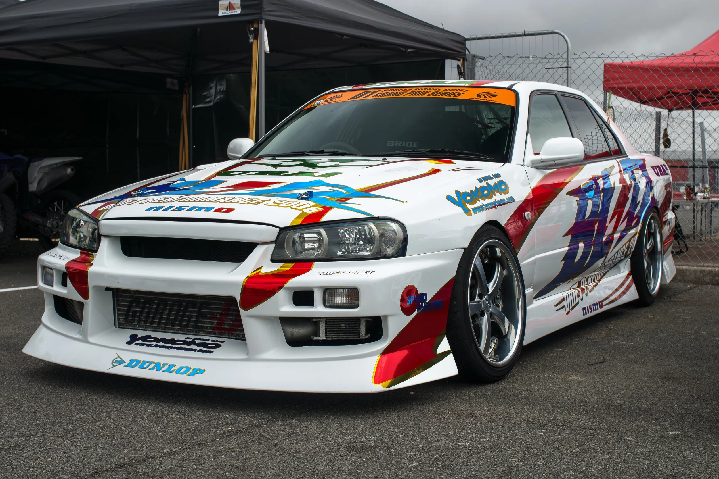White racing car with sponsor decals parked in a paddock area, set against a cloudy sky backdrop.