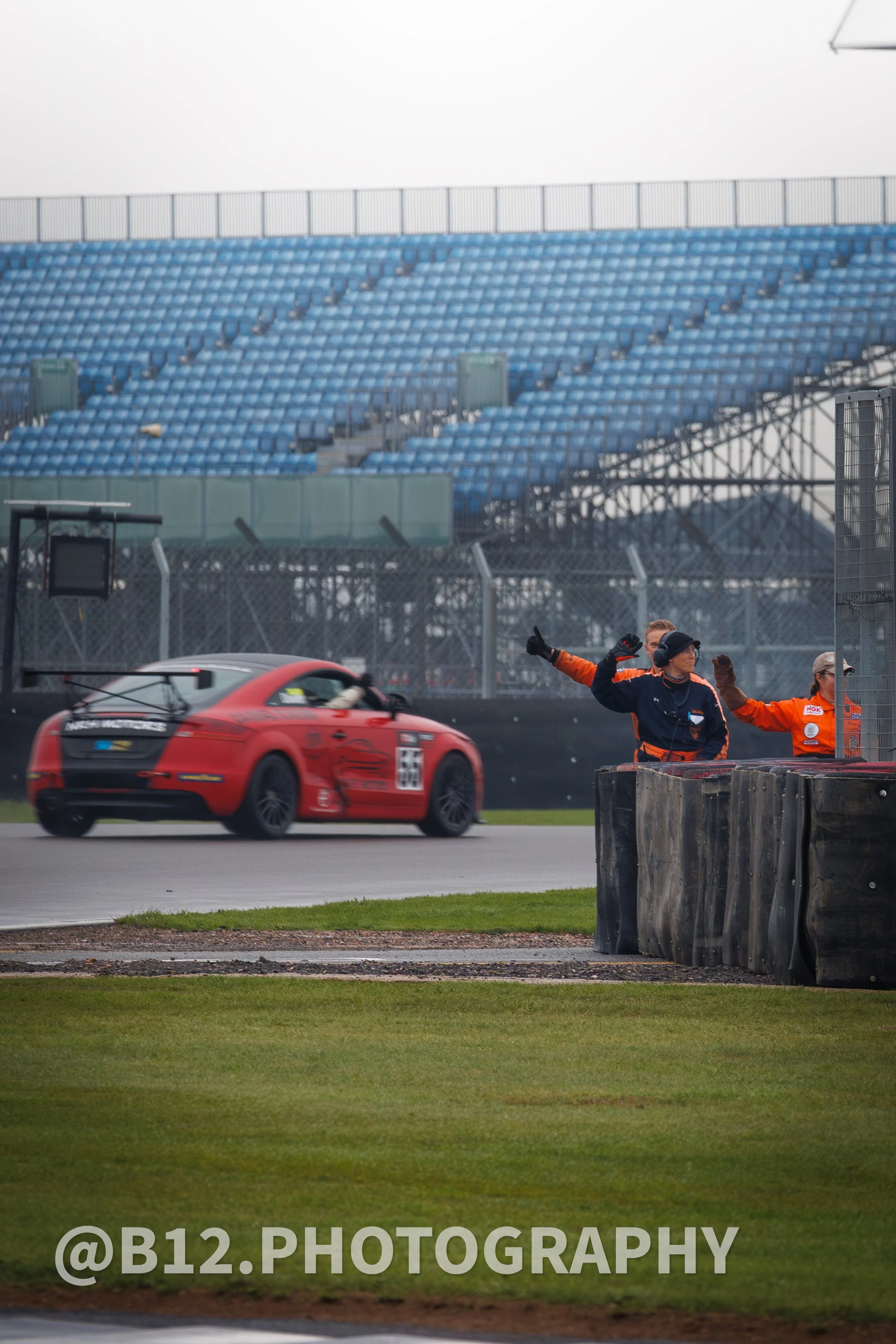 A red race car on a race track with two marshals waving. Empty blue grandstands in the background.