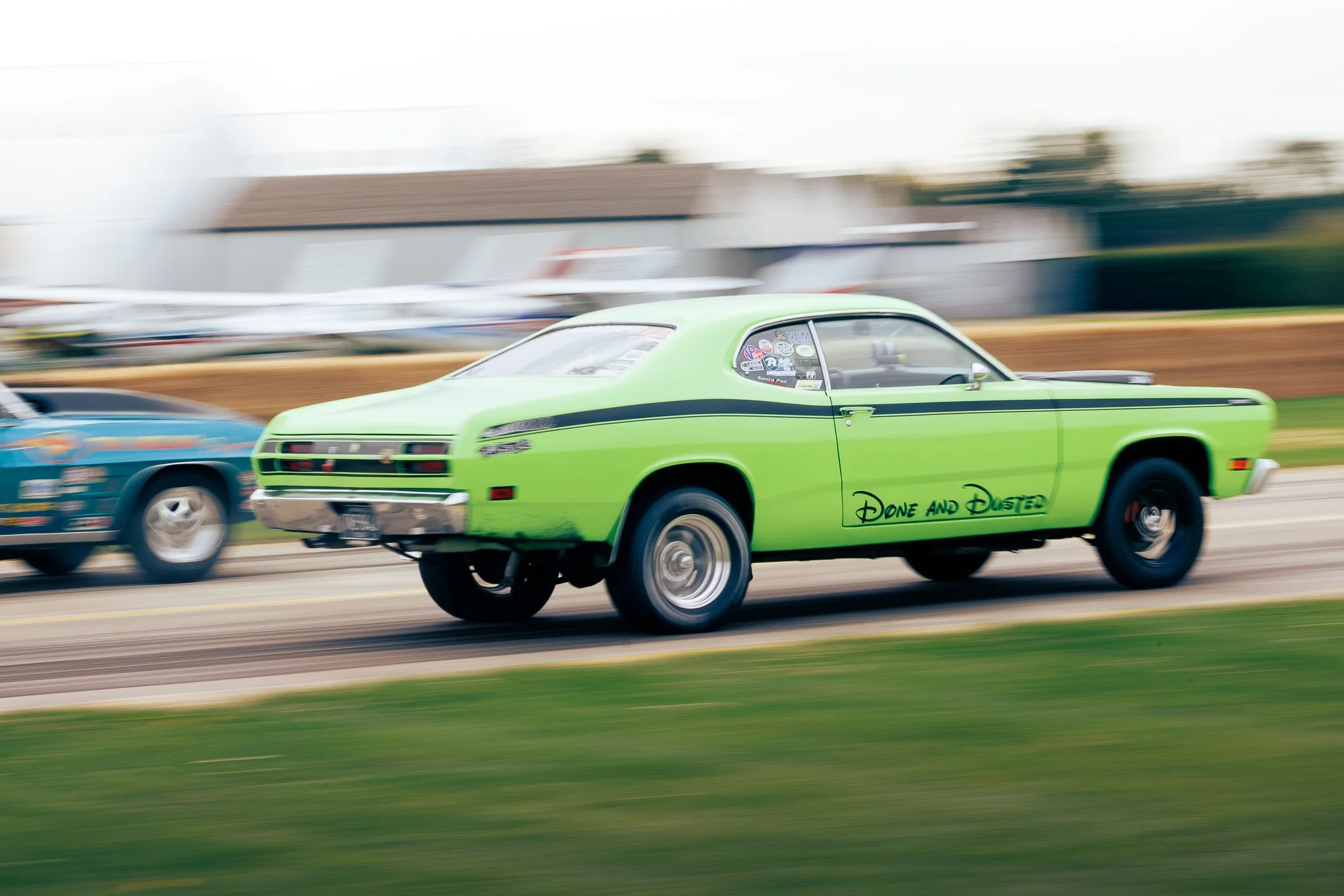 Green race car with the words 'Done and Dusted' on the side, moving fast on a track during a race, with other cars and blurred background.
