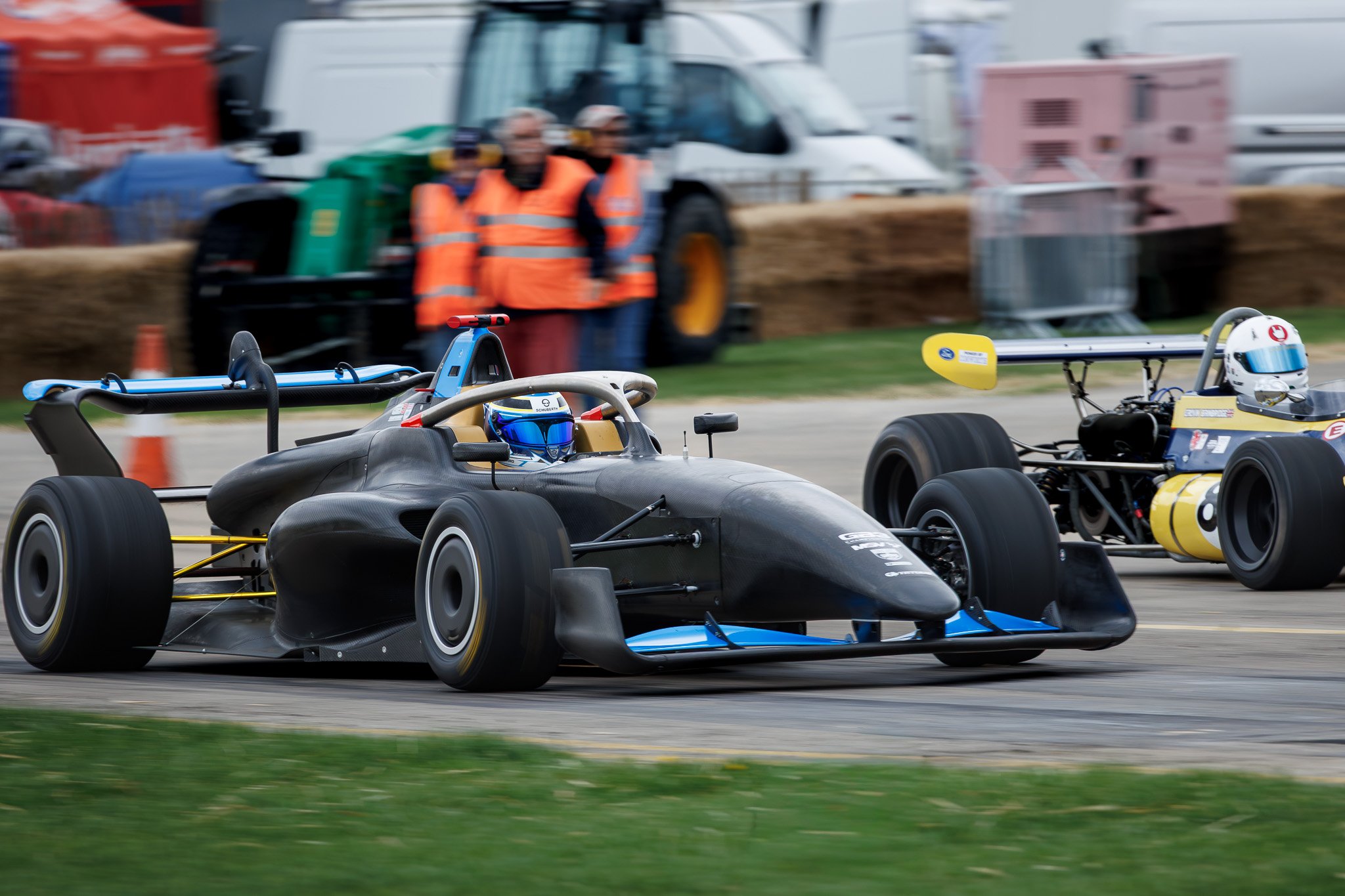 Two open-wheel race cars racing on a track, with safety personnel in orange vests observing in the background.
