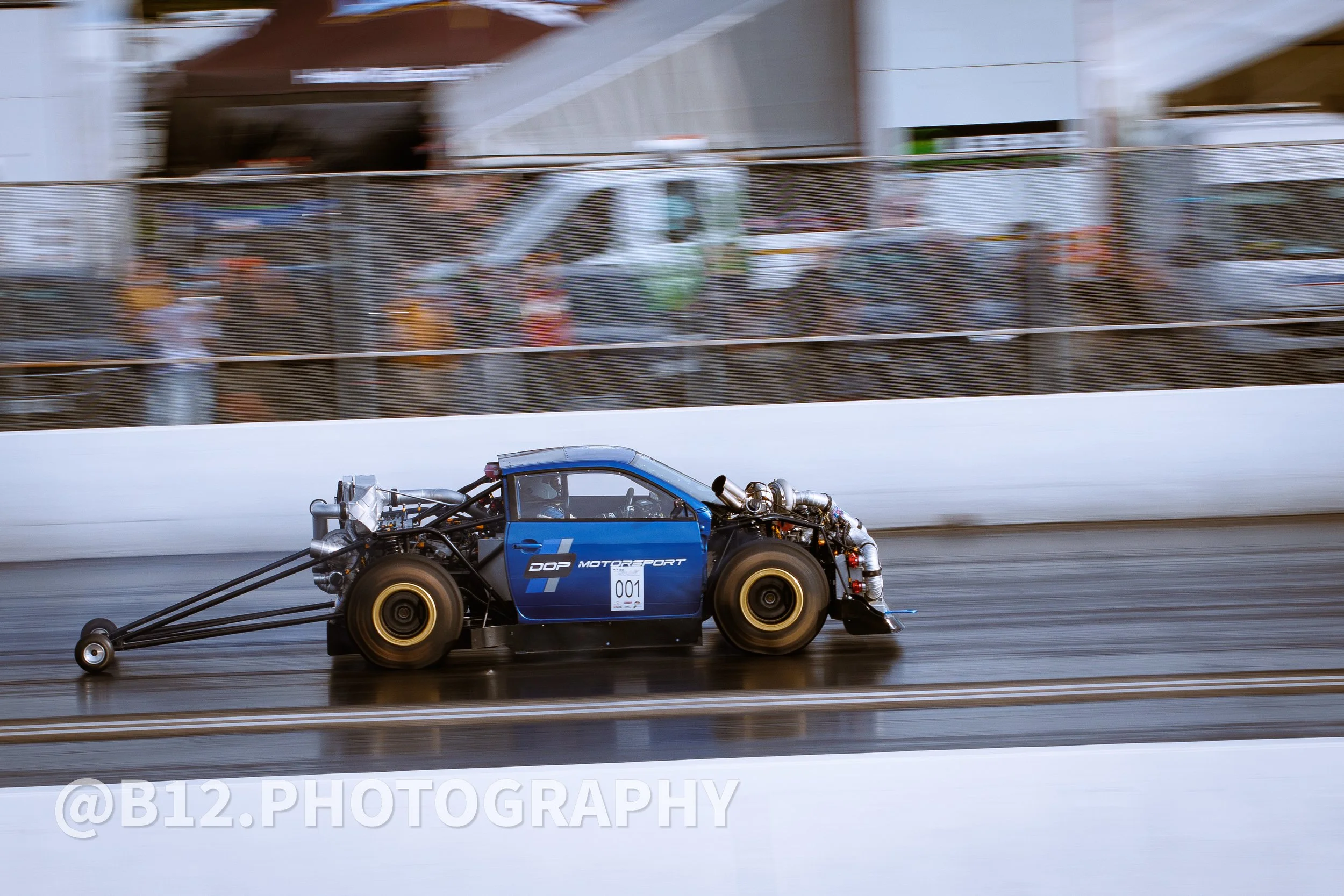 A blue race car with the number 001 on the door is speeding along a racetrack, with a motion-blurred background of spectators and structures.
