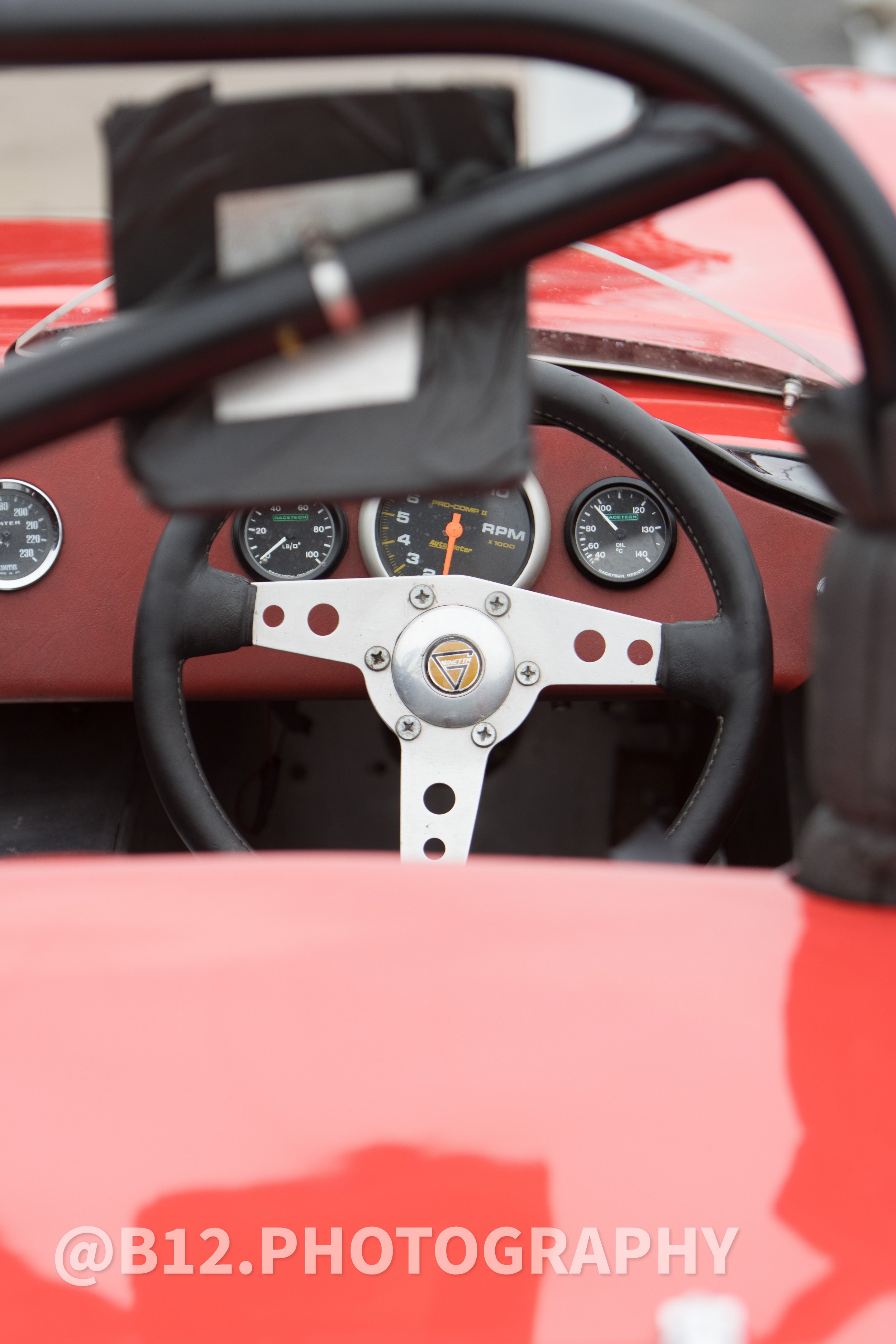 Close-up of a vintage racing car's interior, showing a steering wheel with a platinum-colored center and black rim, dashboard gauges including RPM, speed, and oil temperature, with a red body and a black windshield frame.