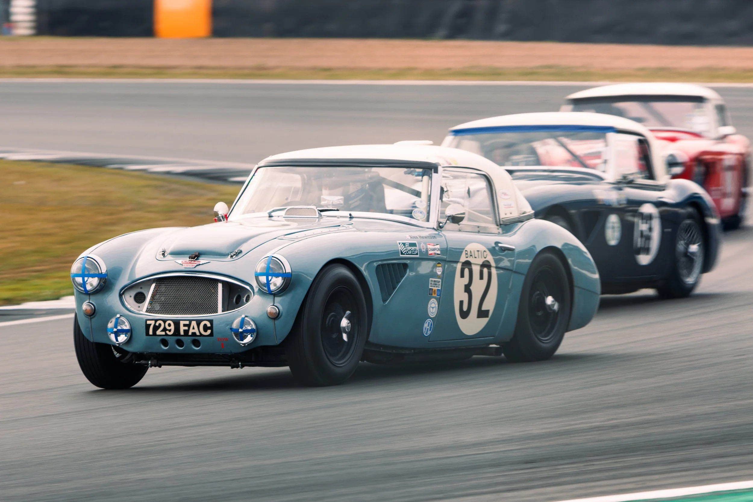 A vintage race car with number 32 on the side, silver-blue color, driven on a racetrack with three other race cars behind it.