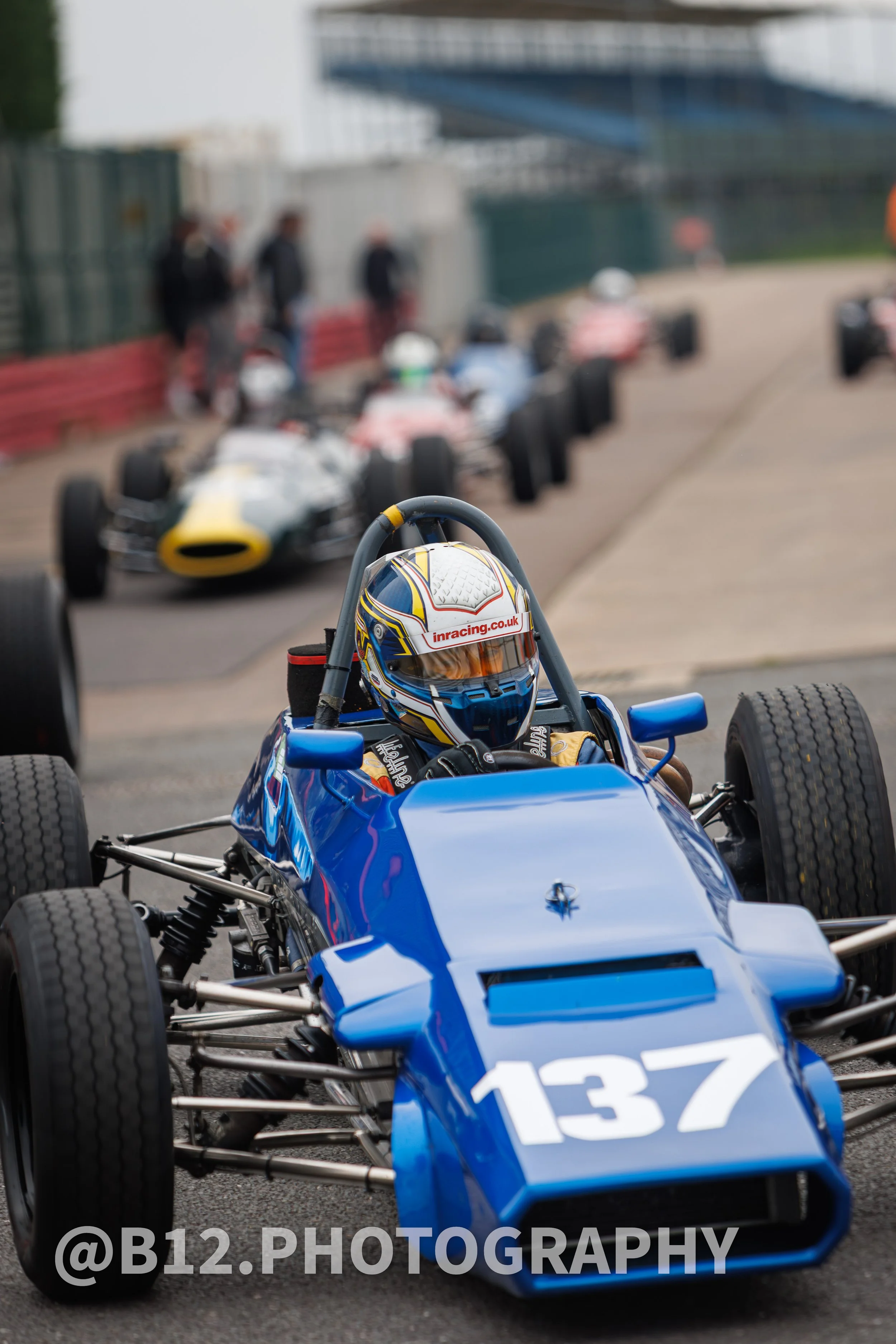 A race car driver sitting in a blue race car with the number 137, on a race track with other race cars and people in the background.