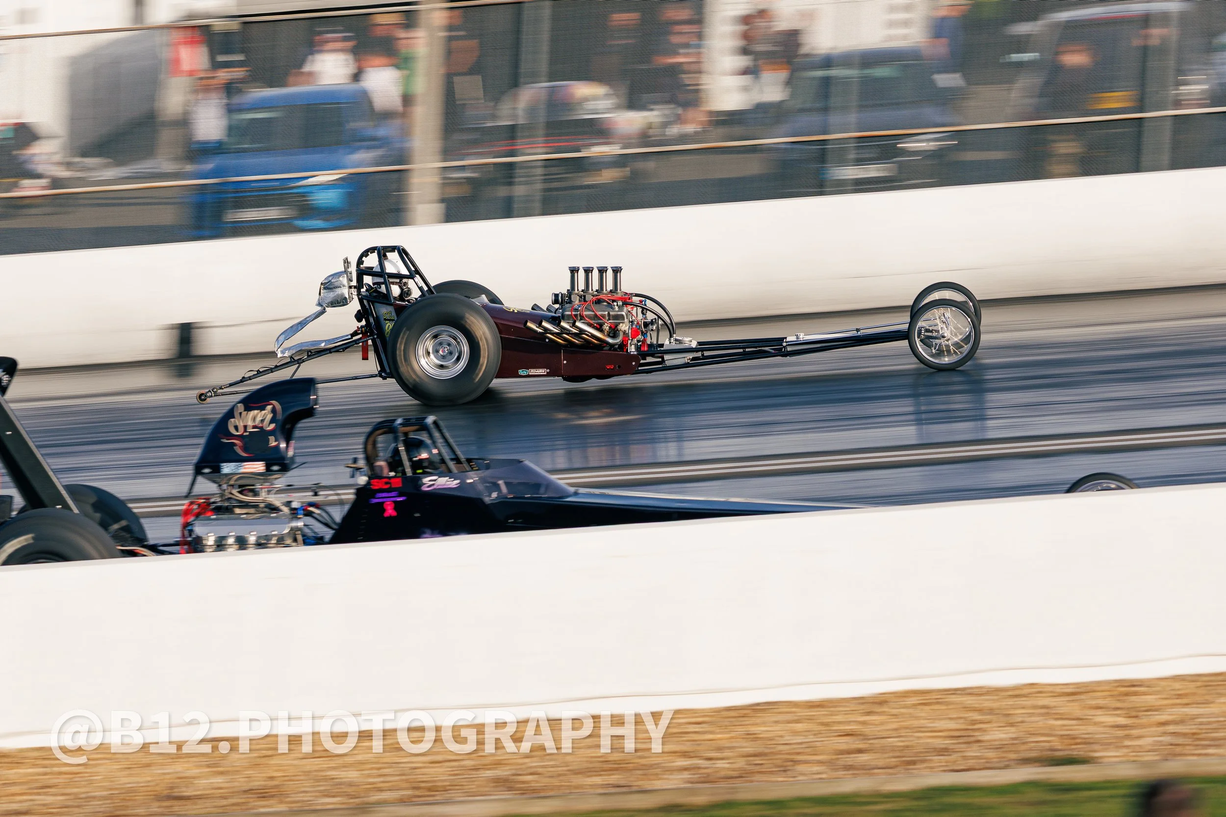 Two drag racing cars speeding down a race track, with spectators blurred in the background. The black car is closer to the viewer, while the maroon car is further ahead.