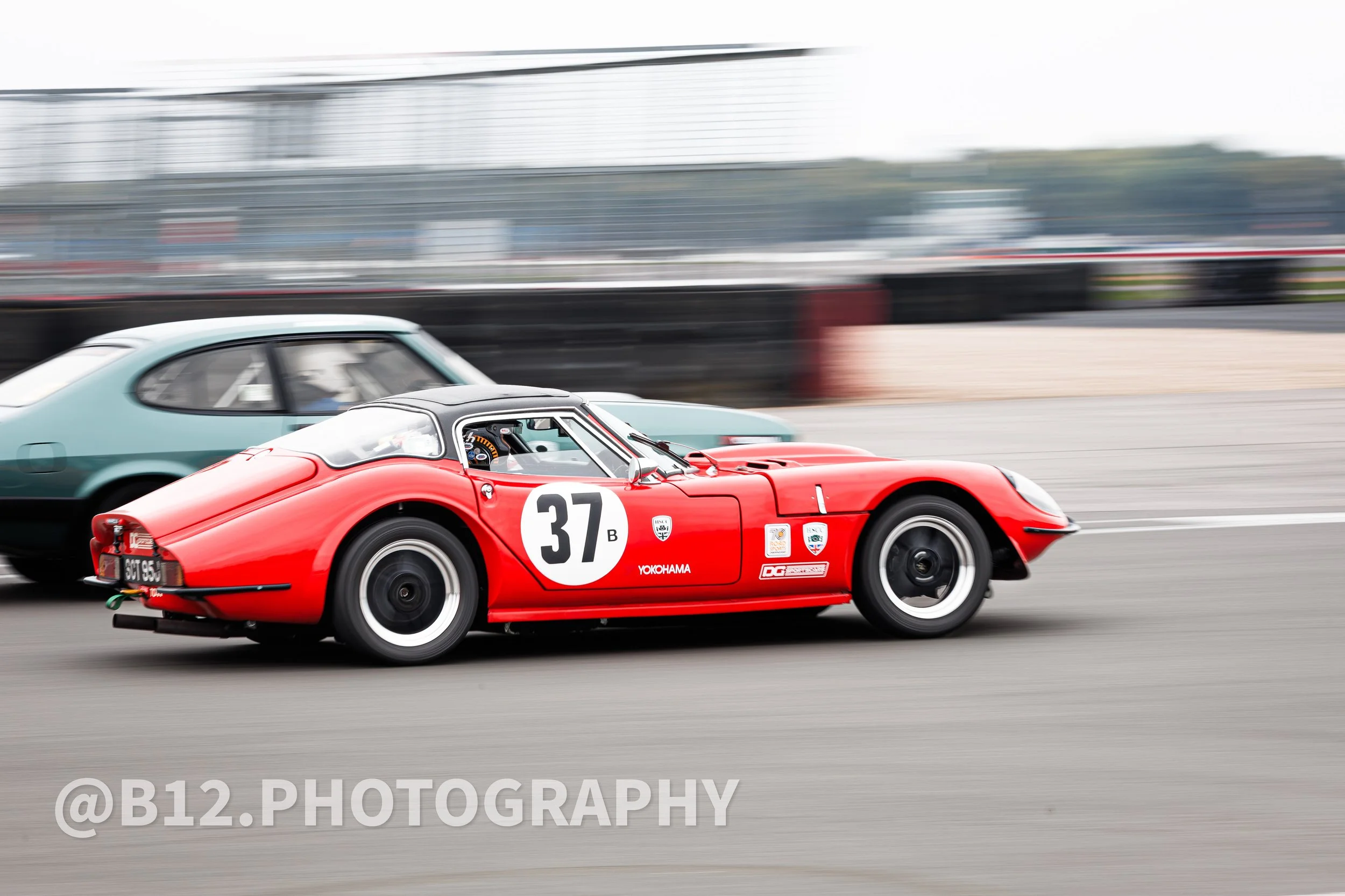 A red vintage race car with the number 37 on the side, driving on a race track, with another car nearby, in motion blur.