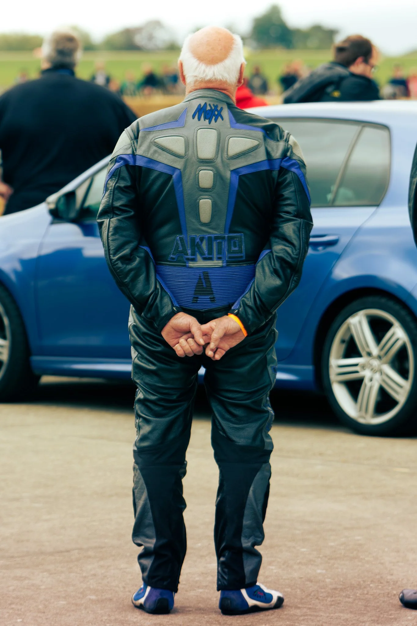 A man in a racing suit stands with his back to the camera, in front of a blue car, in a crowd at a race track or motorsport event.