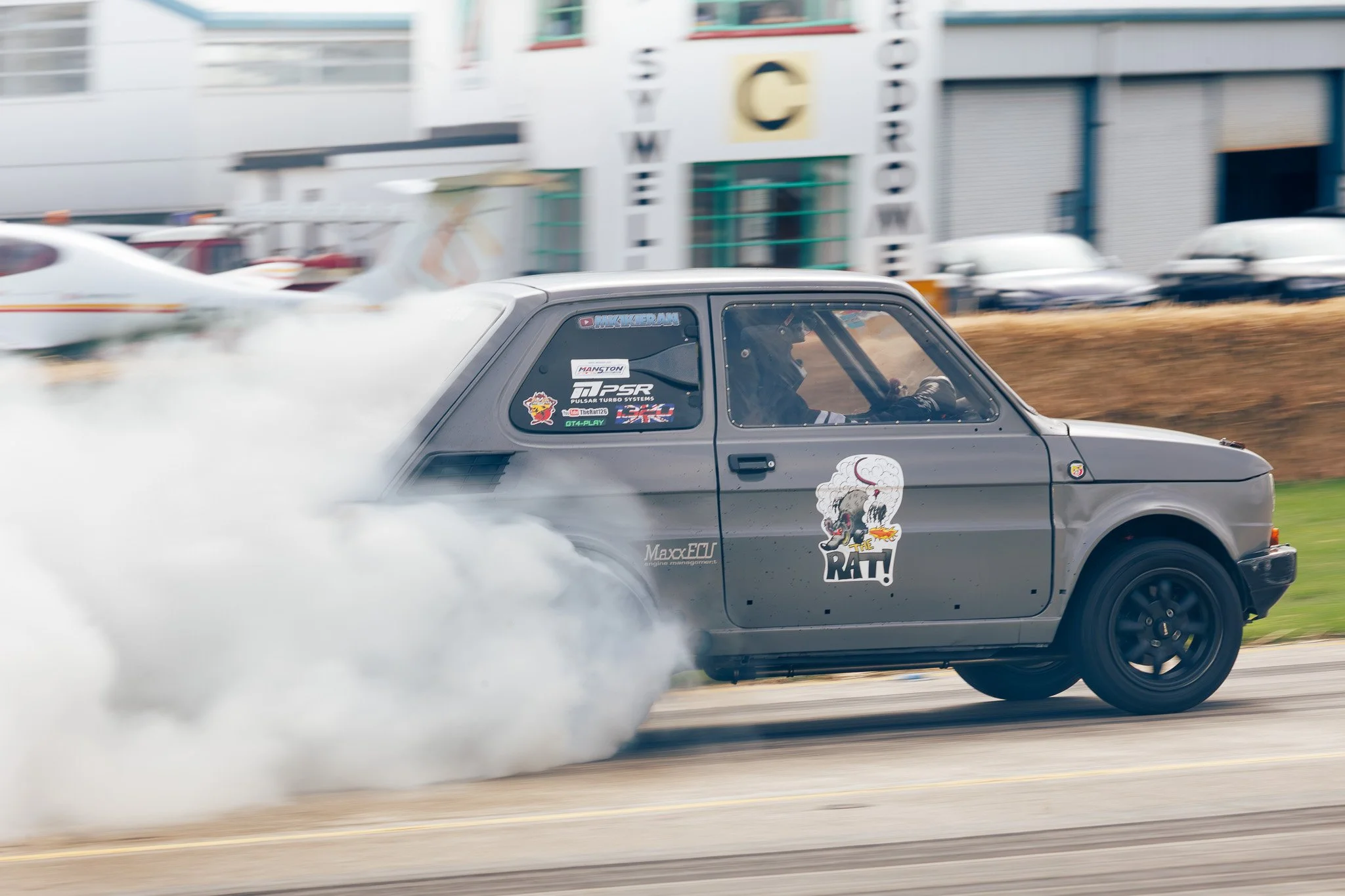 Gray hatchback car performing a smokey burnout on a racetrack, with stickers and graphics on the side, and a building with parked cars in the background.