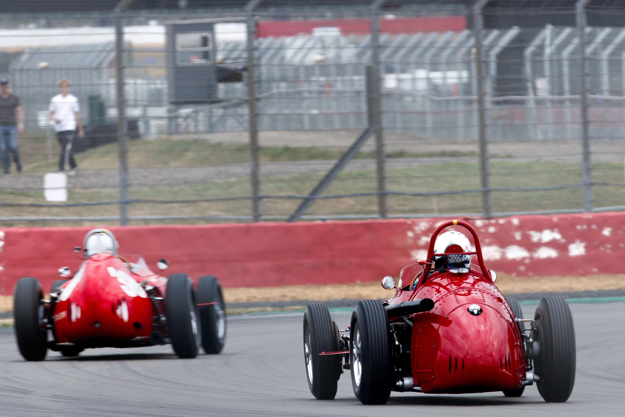Two vintage red race cars on a race track with a concrete barrier and fencing in the background, along with two spectators observing from behind the fence.