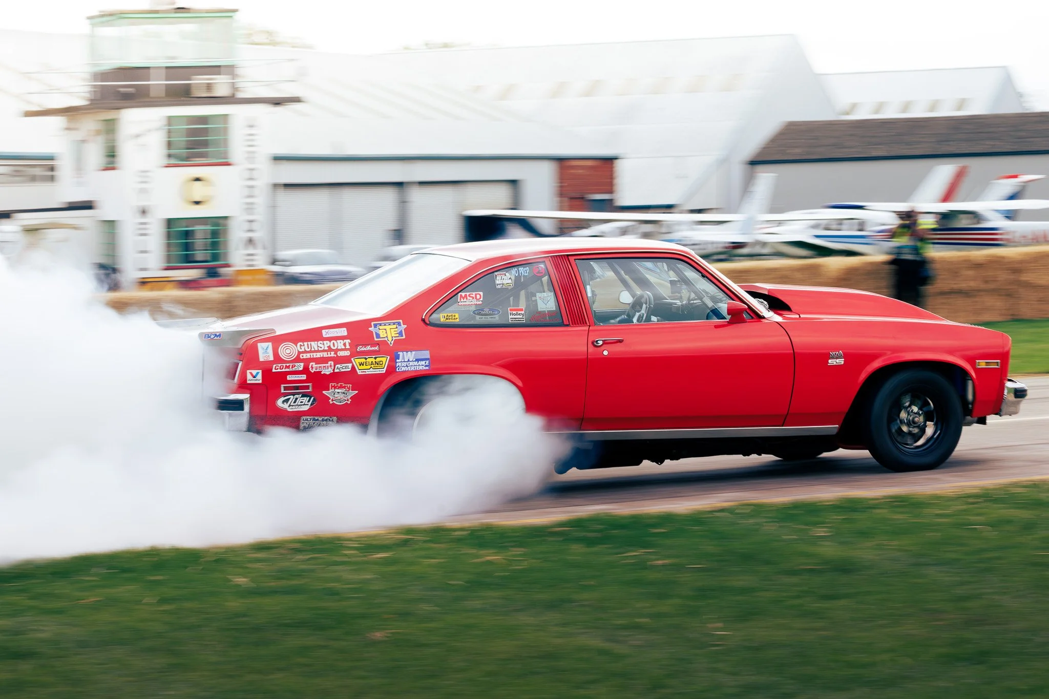A red vintage muscle car performing a burnout at a race track, with smoke billowing from the rear tires and racing stickers on the side.