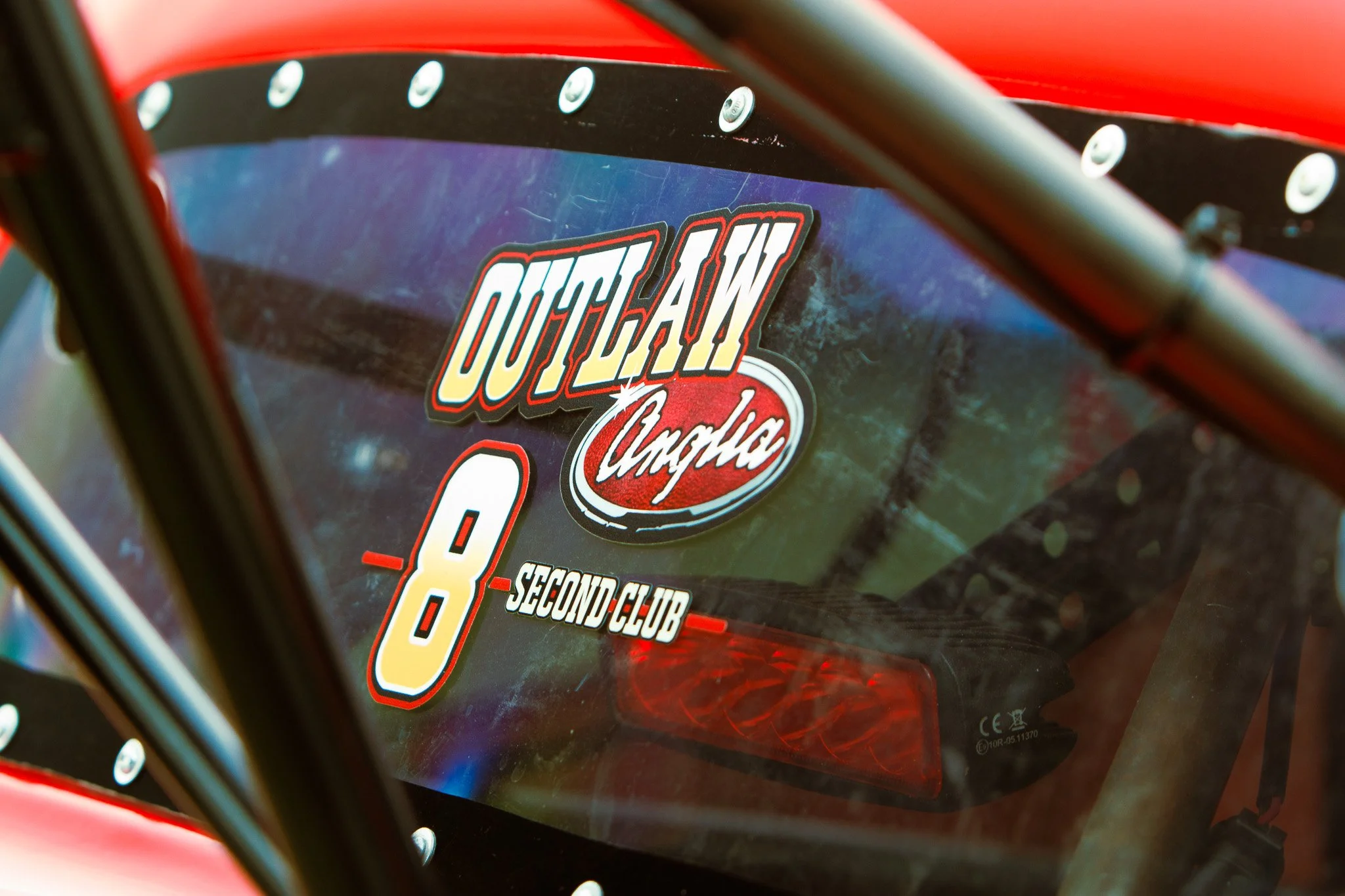 Close-up of a race car window with stickers, including 'Outlaw' and the number 8, with a blurred background of the car interior.