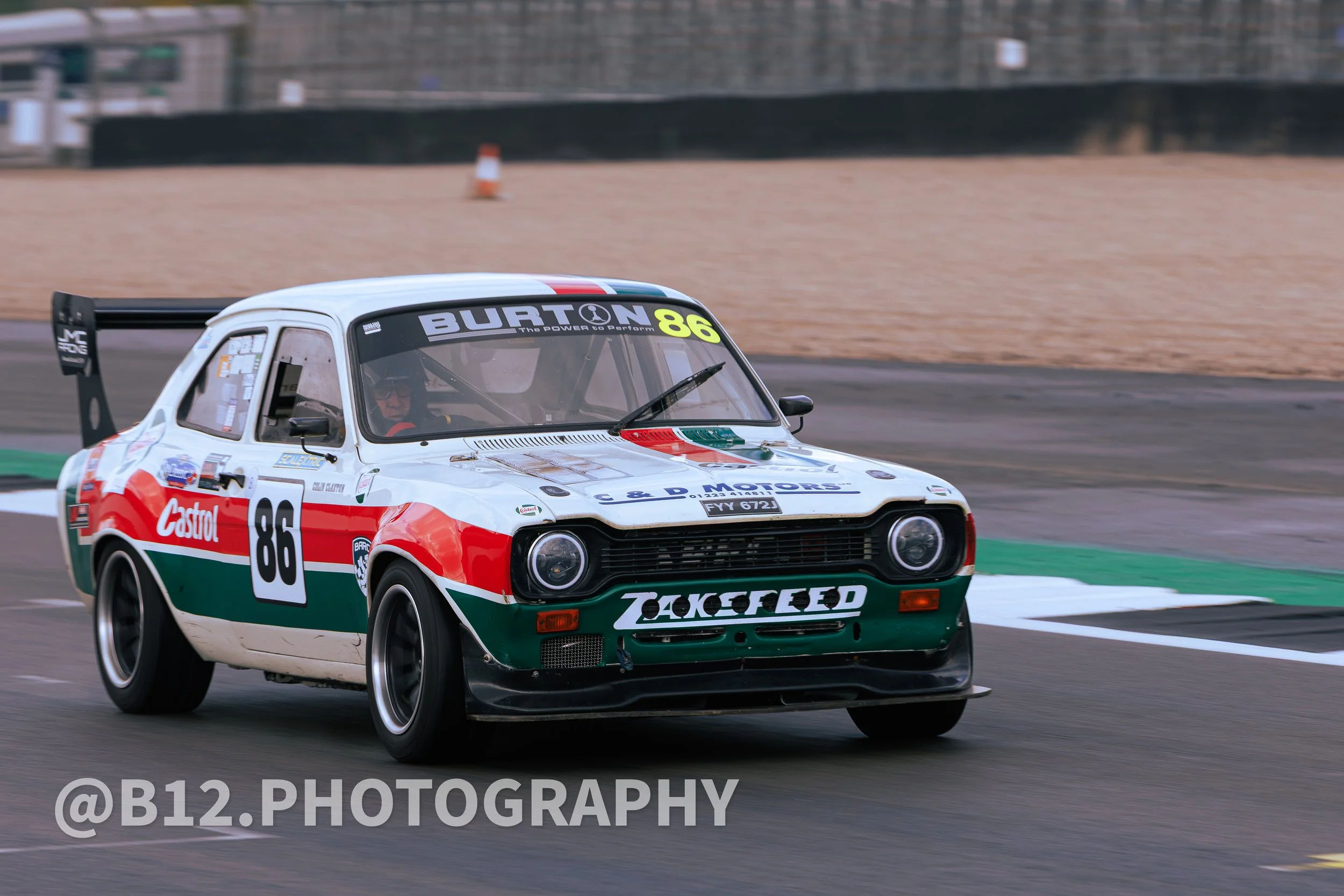 A race car with the number 86, featuring a white, red, and green color scheme with racing decals, is driving on a race track.