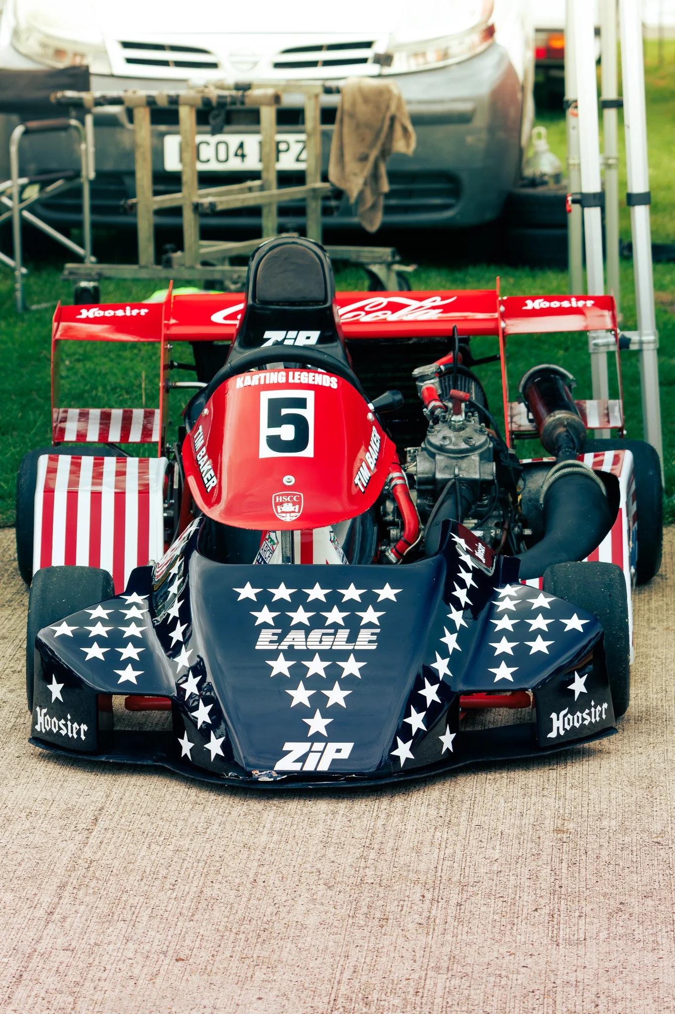 A red and black go-kart with white stars, labeled 'EAGLE' and 'ZIP', parked on a concrete surface in front of a tent and car in a grassy area.