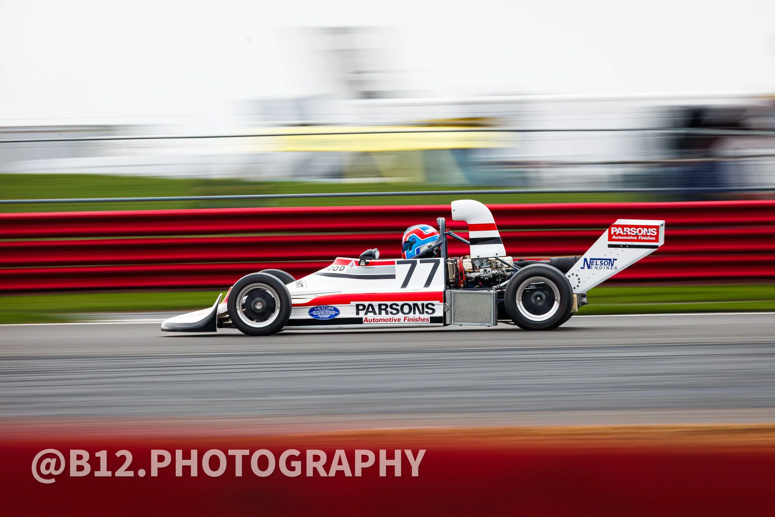 A vintage open-wheel race car with the number 77 and sponsorship logos, speeding on a racetrack with a red safety barrier and blurred background.