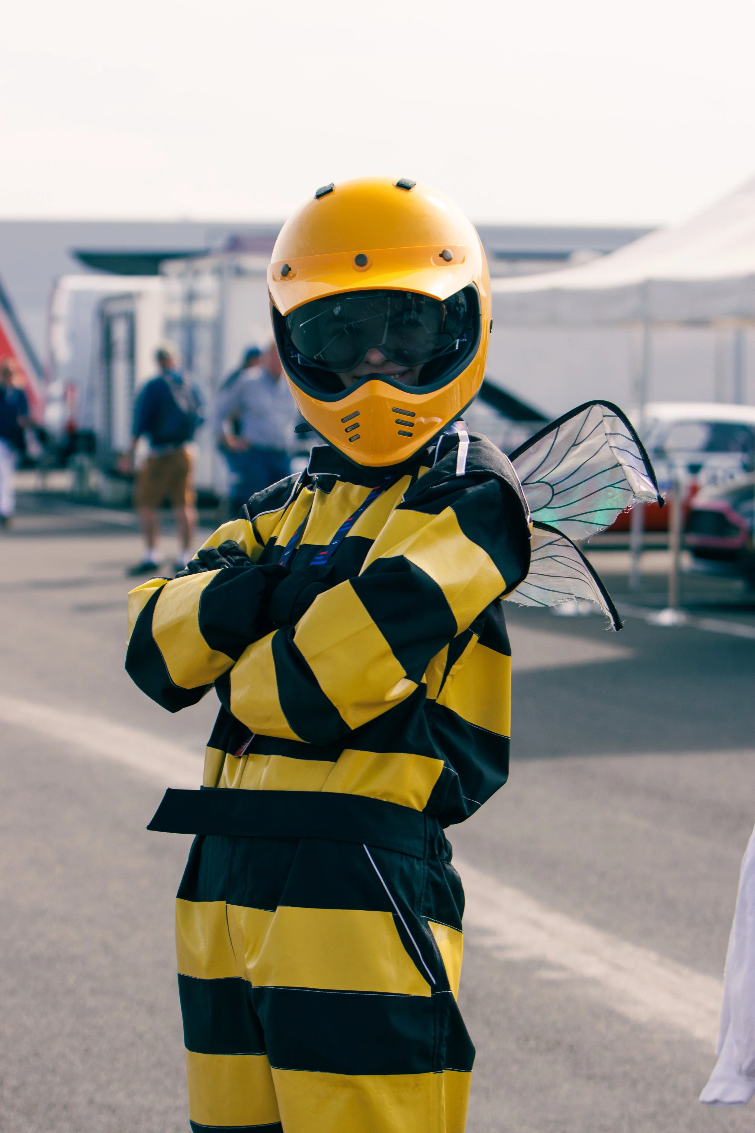 A person dressed as a bee with a yellow helmet, black goggles, yellow and black striped suit, and wings, standing outdoors with arms crossed.