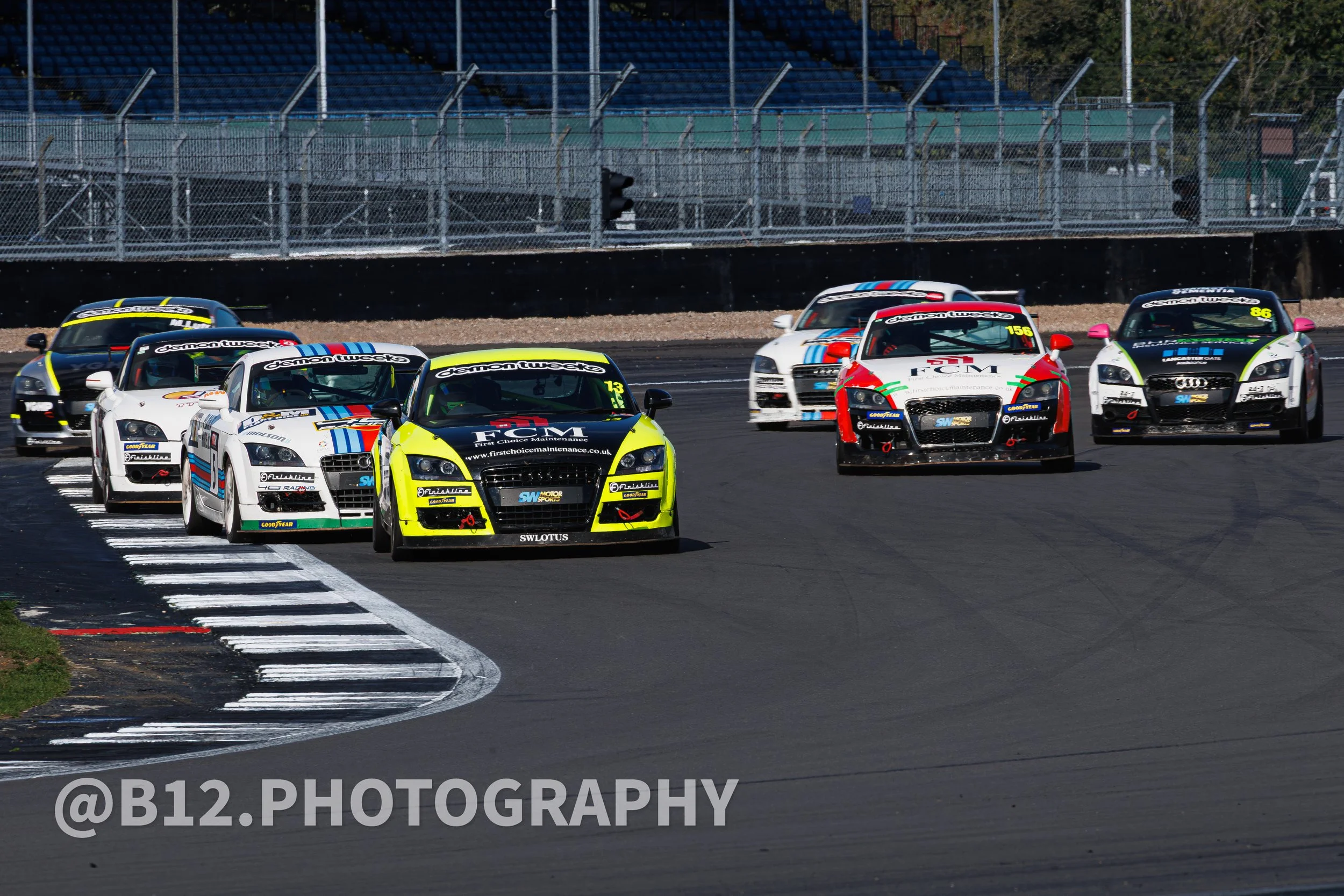 Race cars in a compact formation navigating a curve on a racetrack, with some cars behind others and a fence and seats in the background.